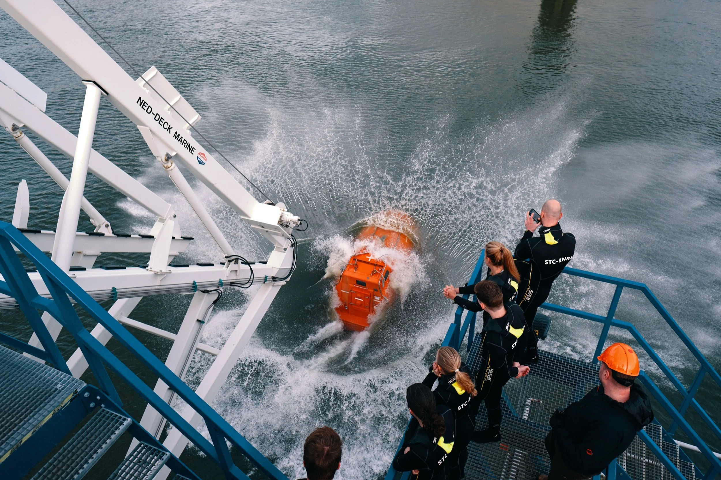 Een reddingsboot met vier mensen op de reling, één persoon springt in het water, terwijl een ander iemand foto's maakt, rondom water met een oranje reddingskrab onder water.