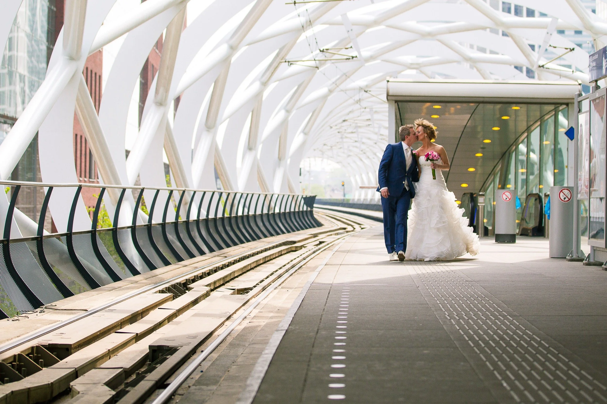 Bruid en bruidegom lopen hand in hand op een moderne tramhalte in een stad, met witte metalen structuur rondom.
