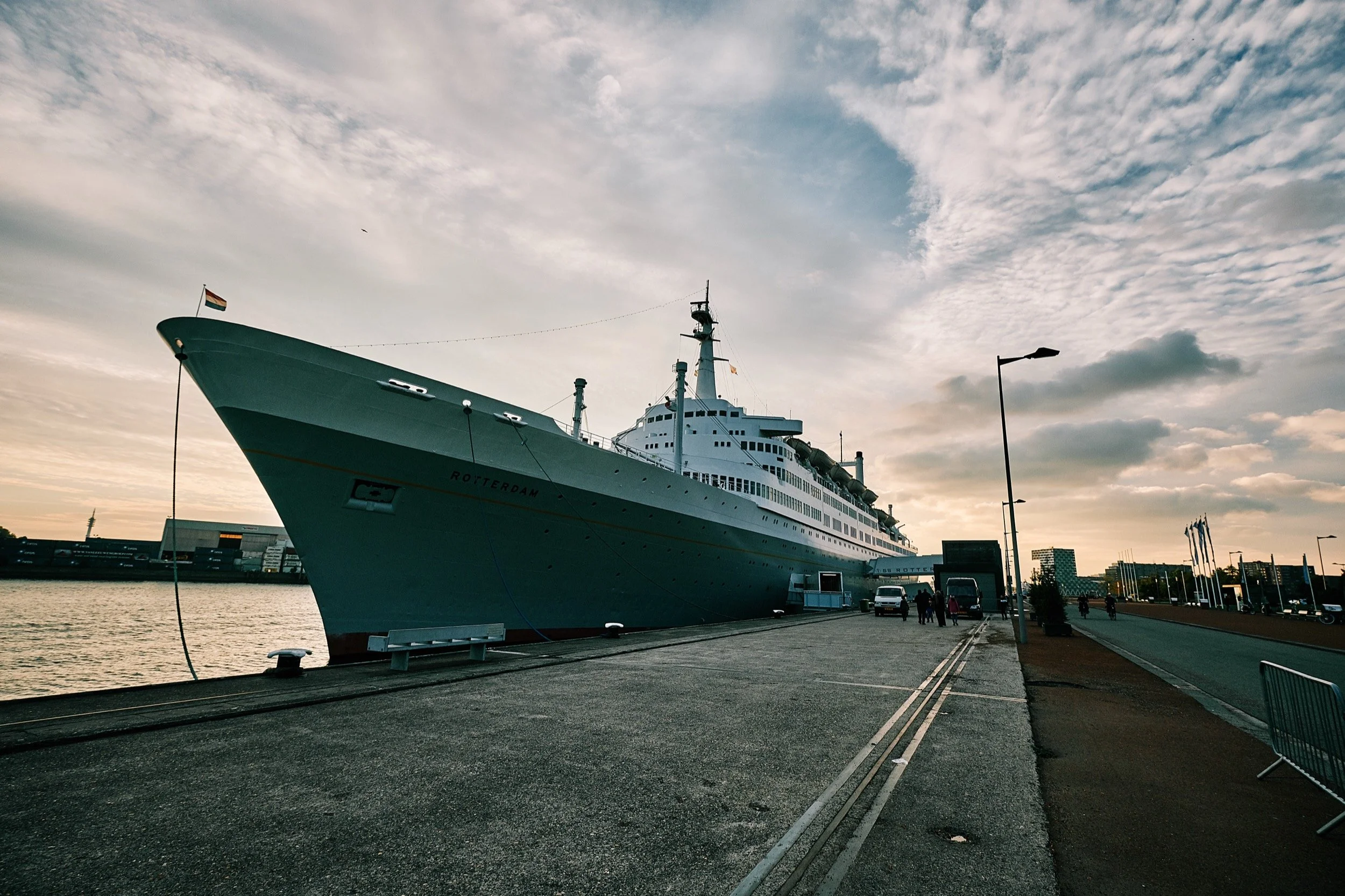 Een grote-cruisevaartuig ligt aangemeerd aan een kade, met een drukke haven en moderne gebouwen op de achtergrond, onder een bewolkte hemel.