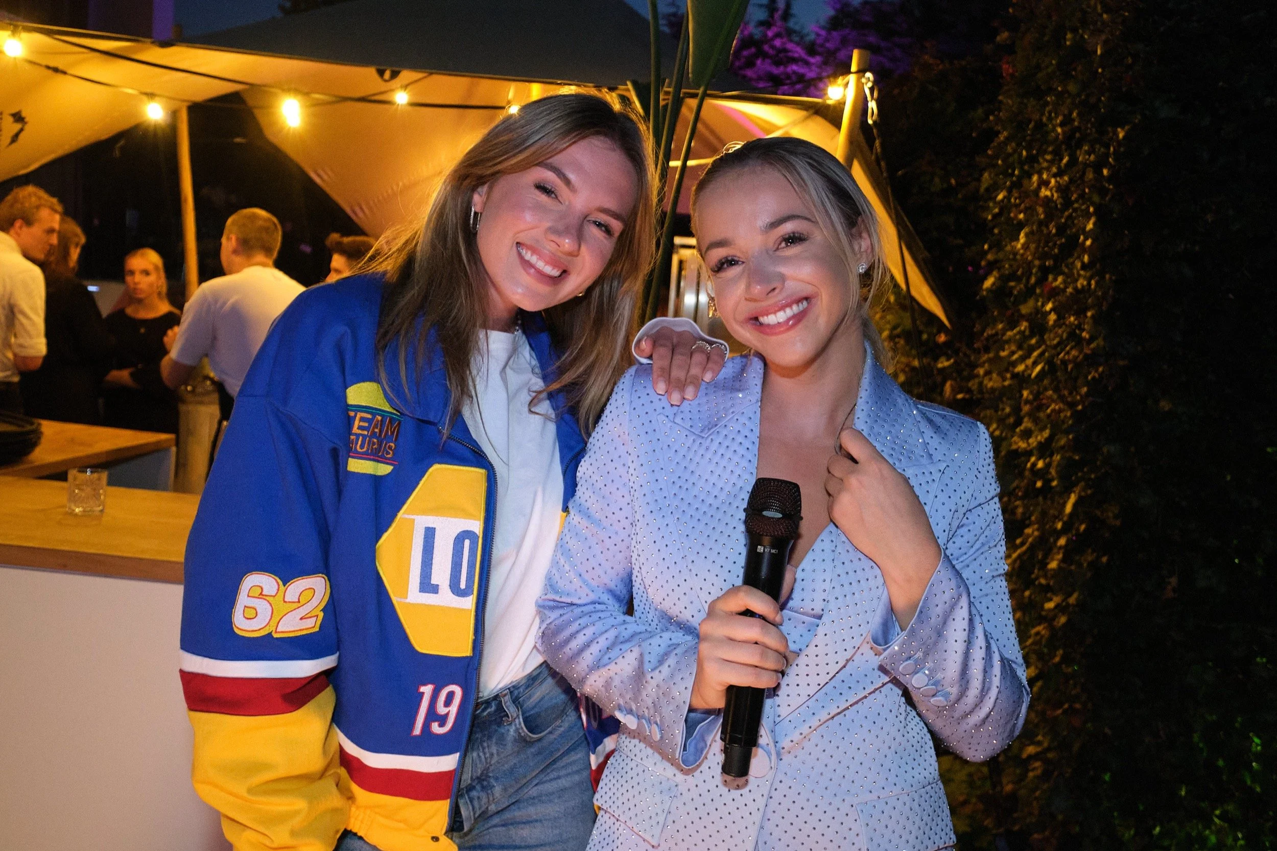 Twee vrouwen glimlachend, één met een microfoon, tijdens een avondbijeenkomst onder een grote parasol met feestverlichting, op de achtergrond andere gasten en groene planten.