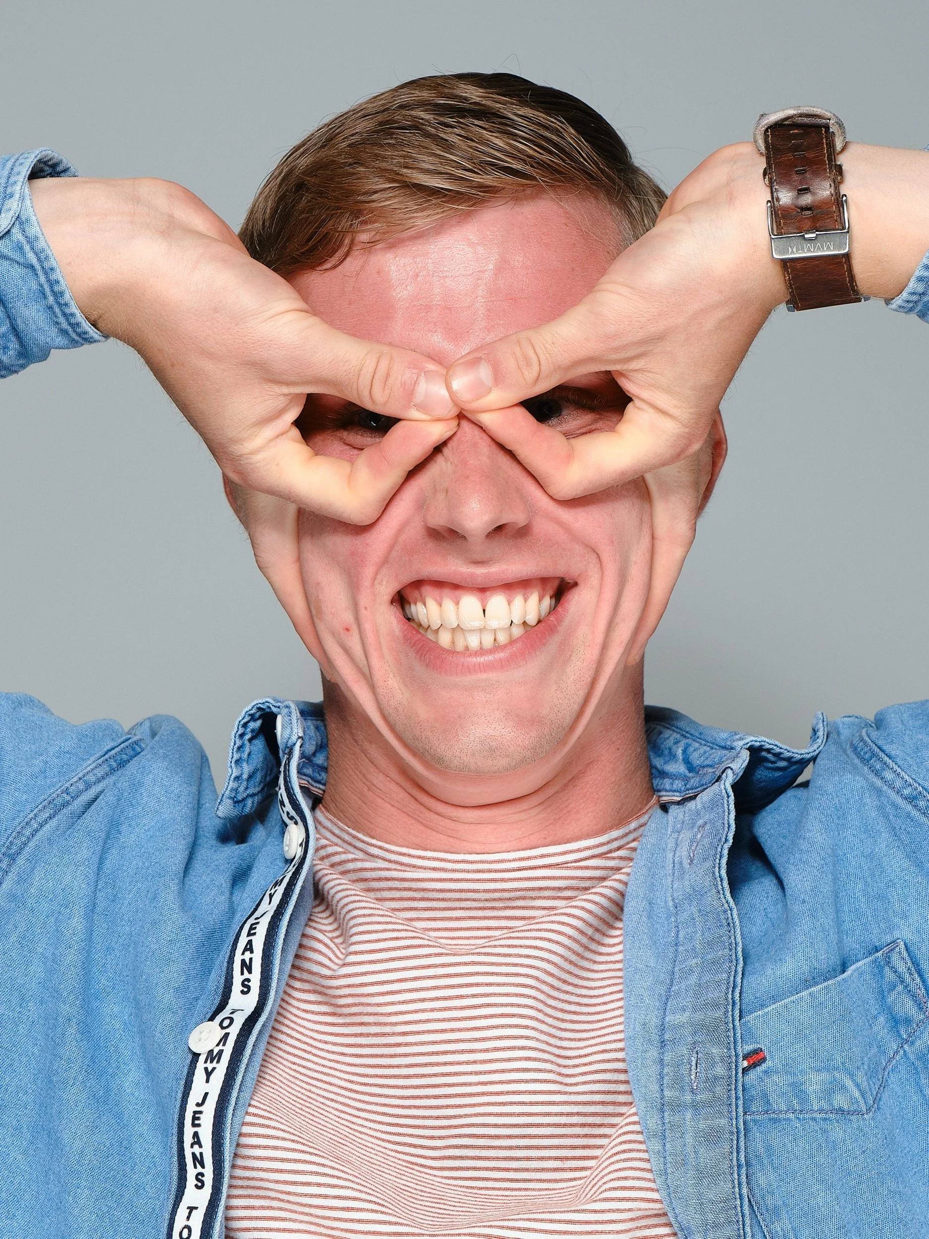 Man die lachend en speels kijkt terwijl hij zijn ogen bedekt met zijn vingers in een hartvorm, gekleed in een denim jas en een gestreept t-shirt.