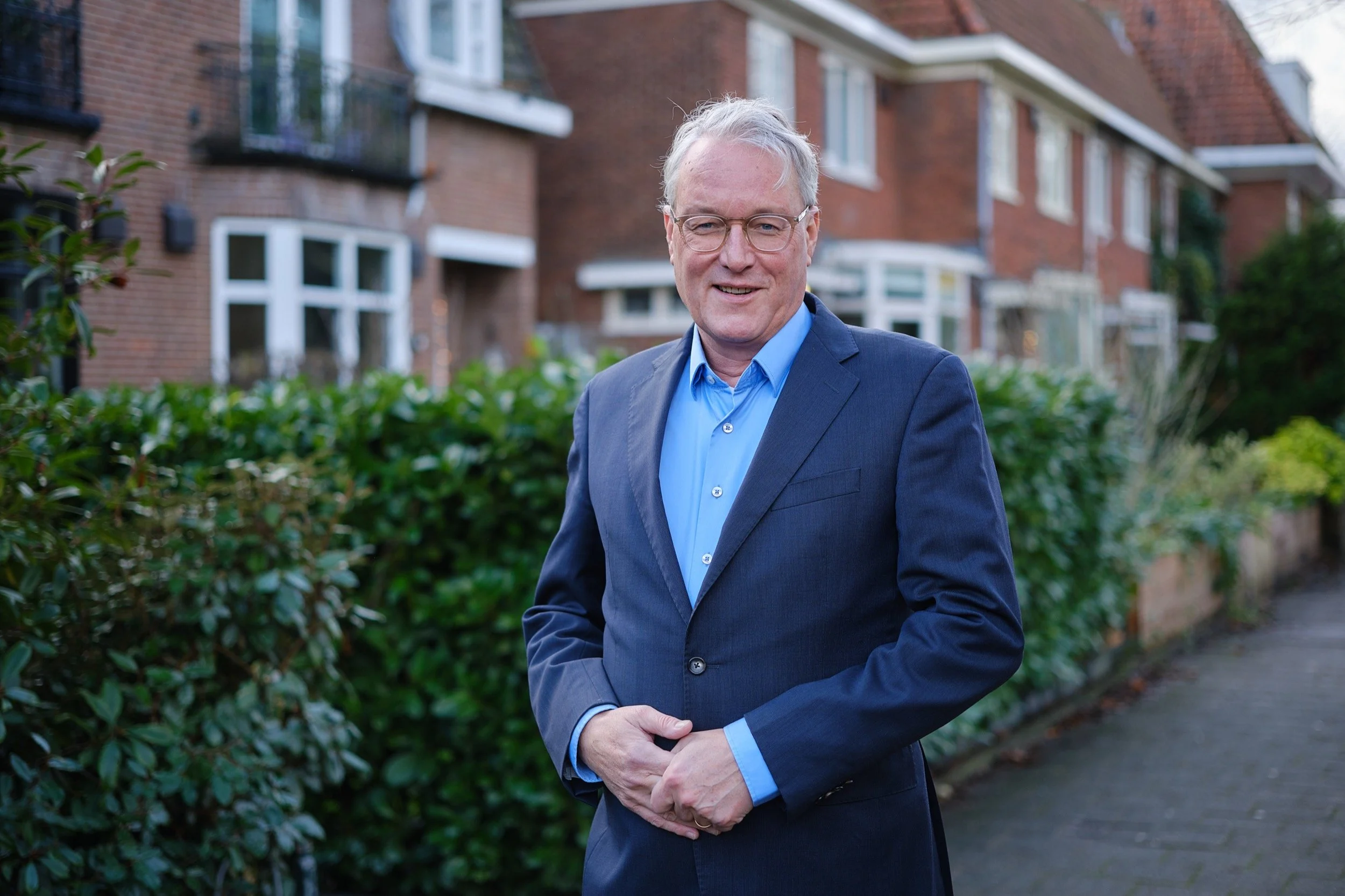 Een oudere man in een blauwe nette jas en bril staat buiten op een straat, met een groene struik en huizen op de achtergrond.