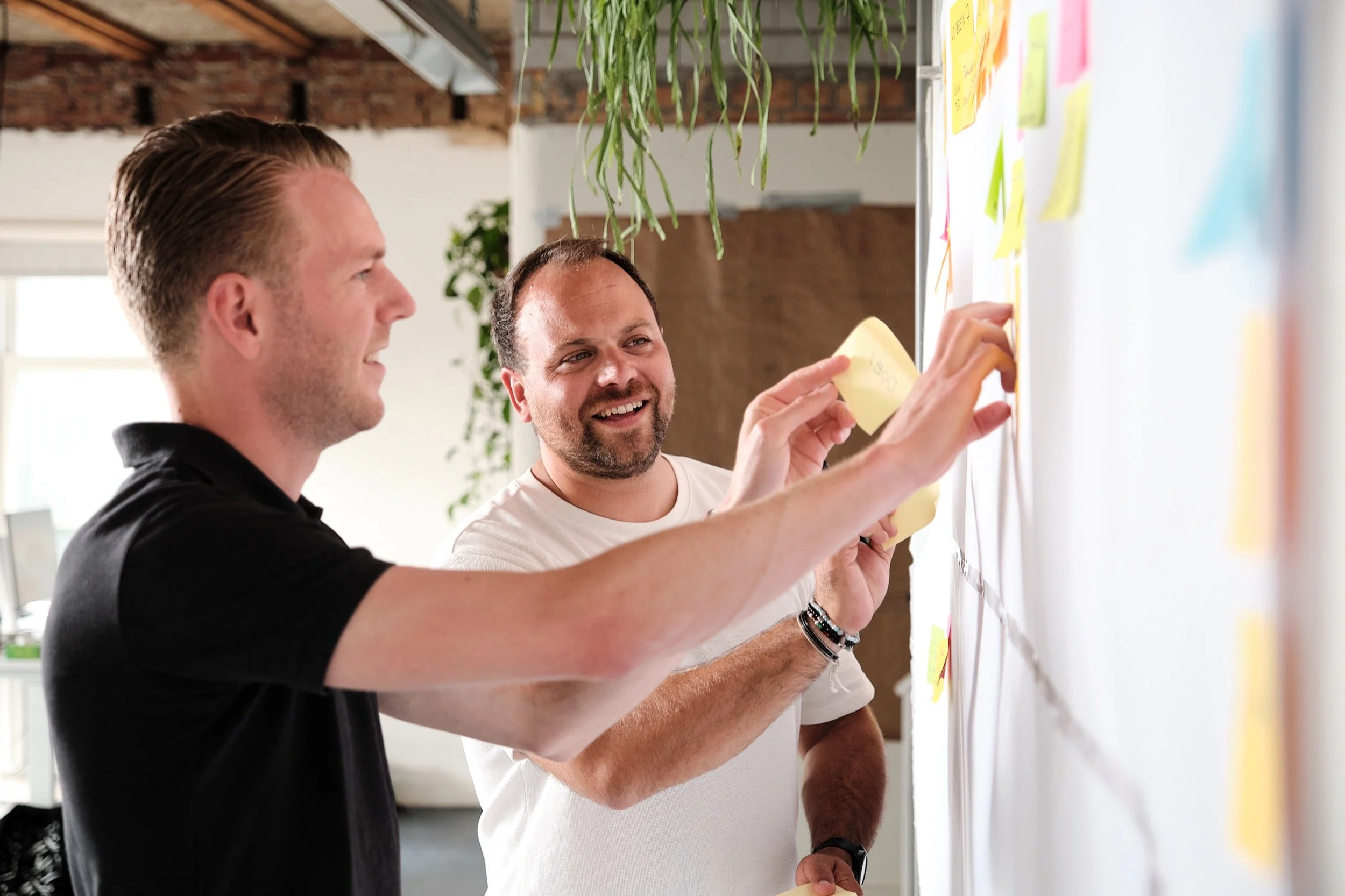 Twee mannen die samenwerken en ideeën uitwisselen met behulp van gekleurde notities op een whiteboard in een kantoor.