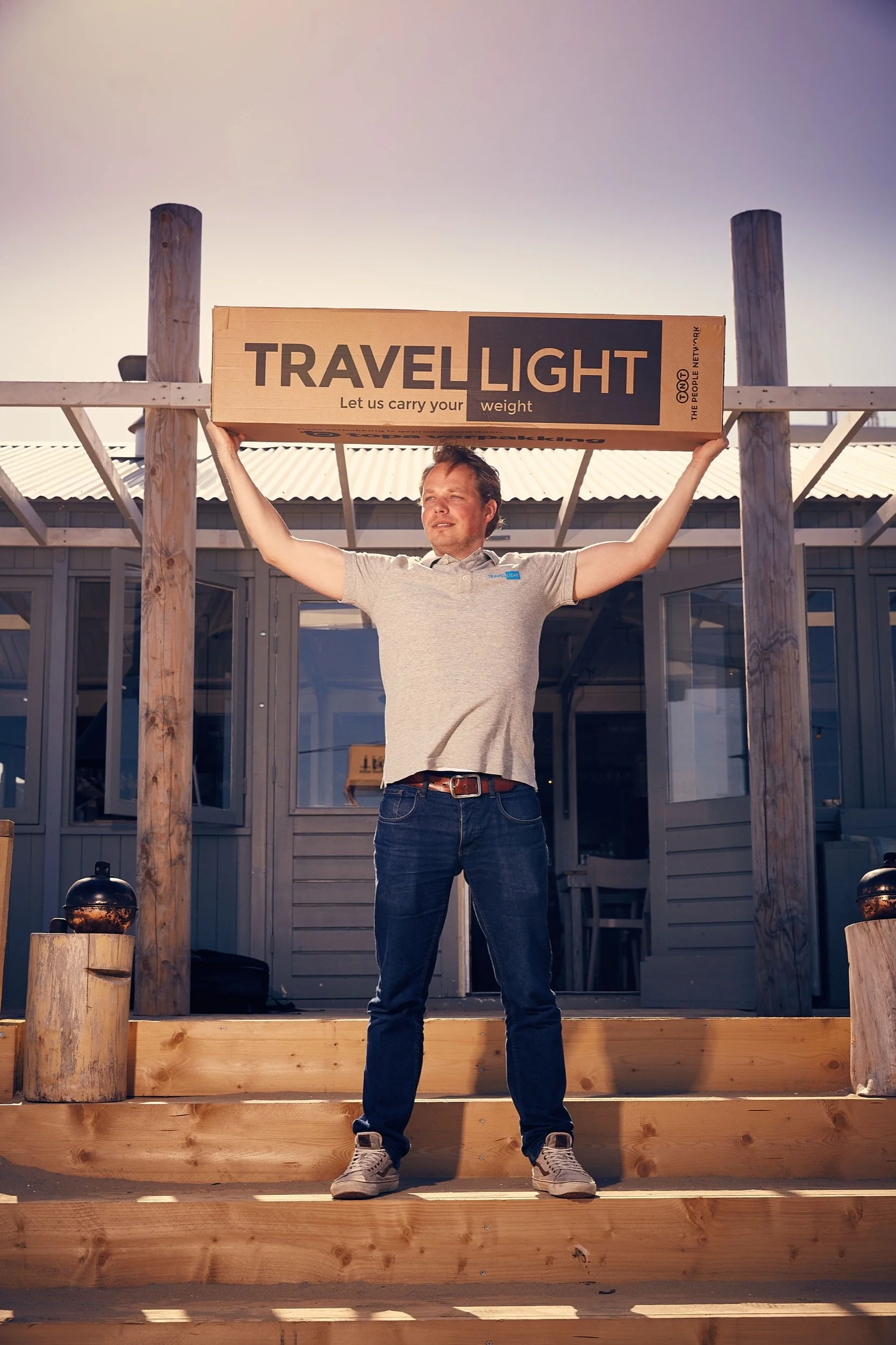 Man staat op houten trappen en houdt een groot kartonnen doos boven zijn hoofd met de tekst 'TRAVEL LIGHT'.