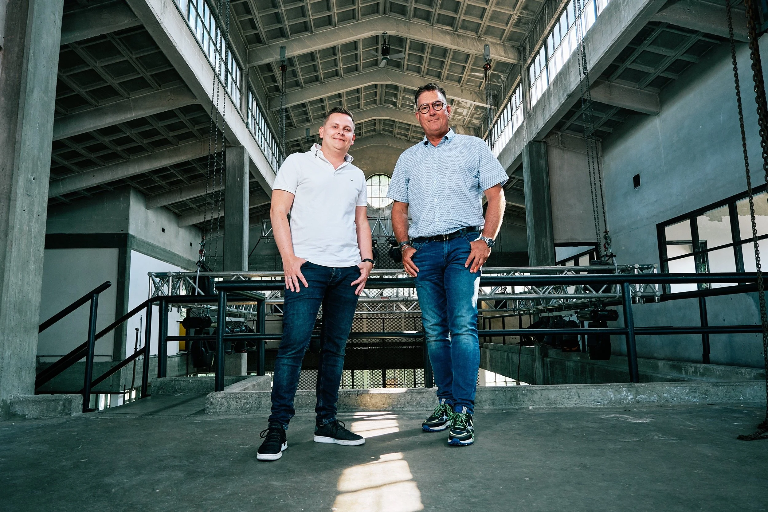 Twee mannen staan binnen in een industriële ruimte met grote ramen en betonstructuren, poseren voor de camera.