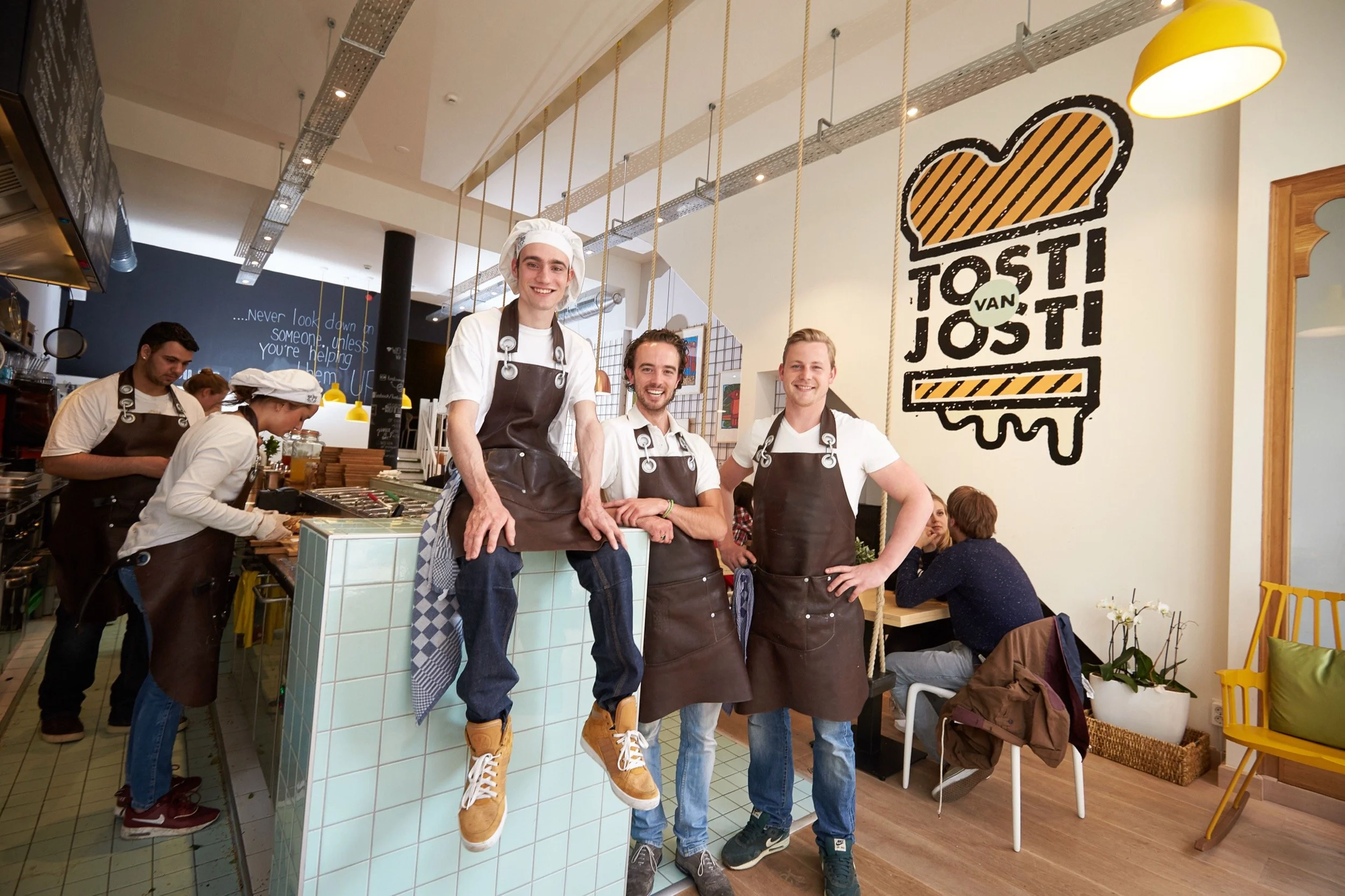 Vijf koks in een restaurant keuken, lachend en in uniform, met een grote muurkunstwerk dat zegt 'Tosti van Jost' met een tosti-illustratie, in een gezellige eetgelegenheid.