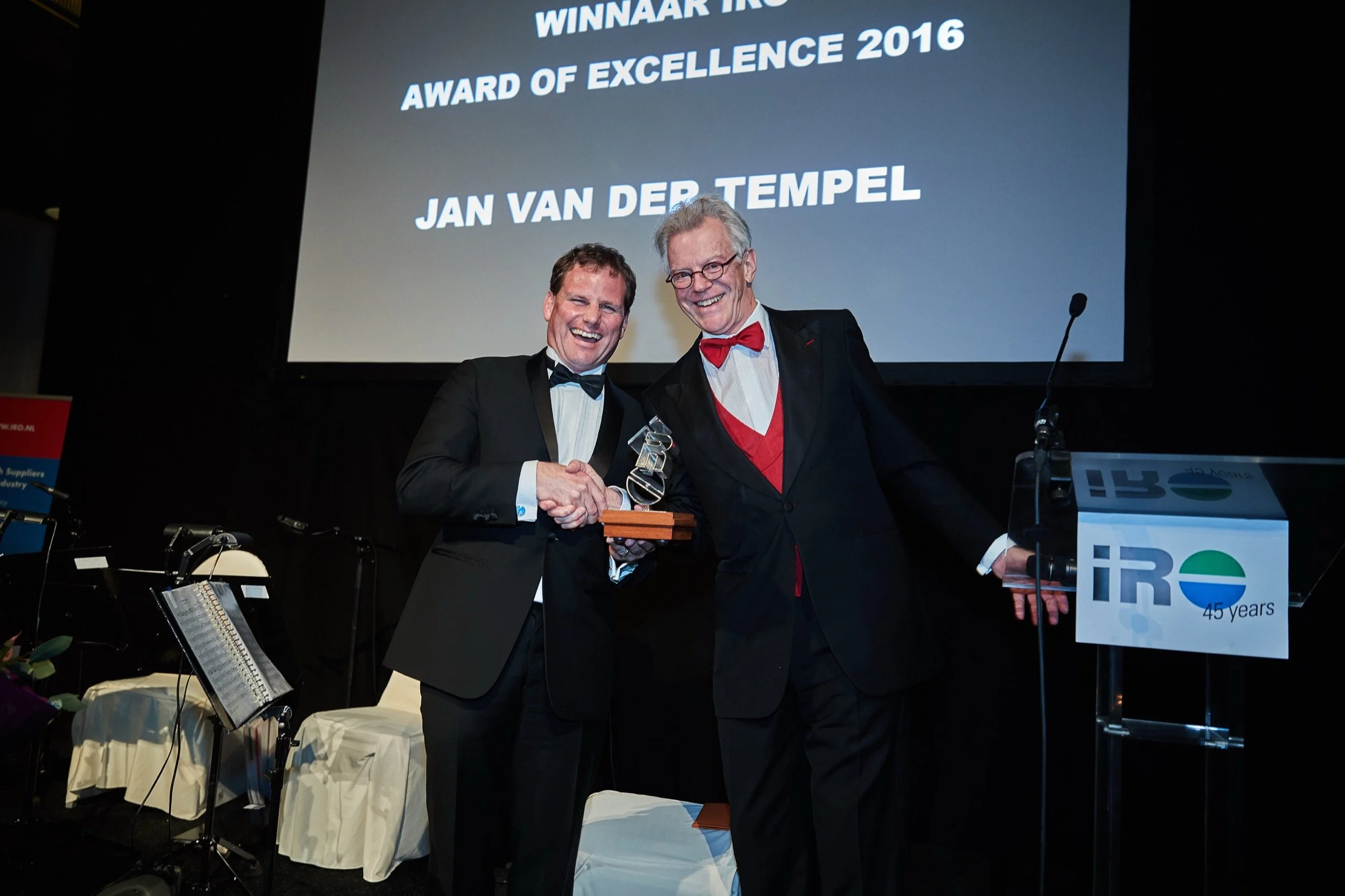 Twee mannen in formele kleding glimlachen en schudden handen tijdens een awarduitreiking. Op de achtergrond een groot scherm met tekst: 'WINNAAR TRO', 'AWARD OF EXCELLENCE 2016', 'JAN VAN DER TEMPEL'.