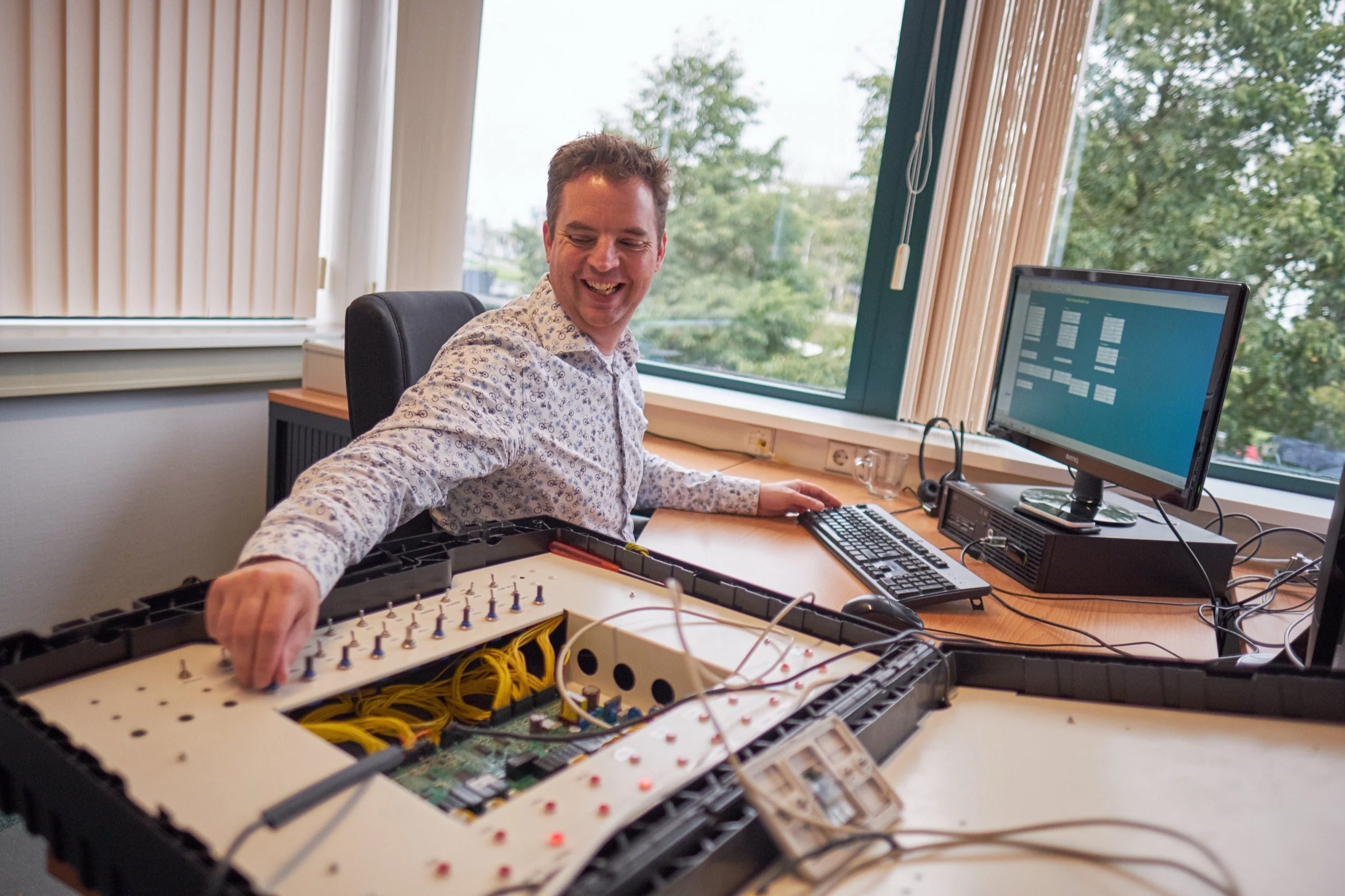 Man in een kantoor met open elektrische kast en computer, lachend en werkend aan technische apparatuur.