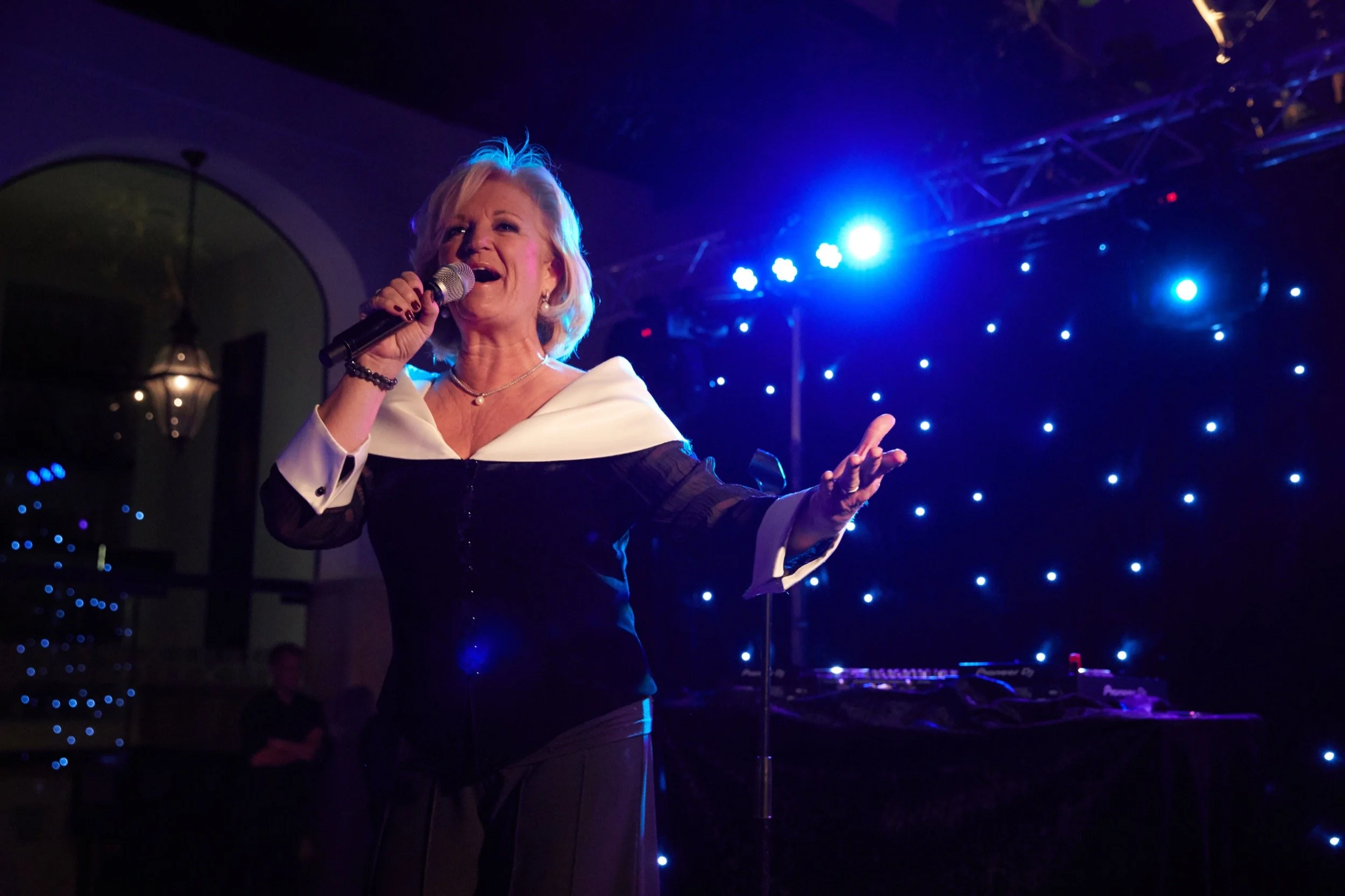 Vrouw zingt met microphone op een podium, blauwe achtergrondverlichting en sterrenlichtpatroon.
