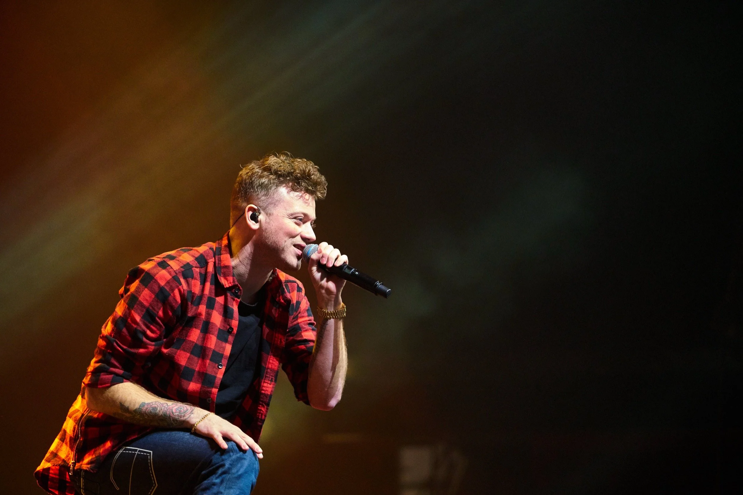 Een man zingt tijdens een concert, gekleed in een rood en zwart geruit overhemd, met een microfoon in de hand en een tattoo op zijn arm, tegen een donkere achtergrond.
