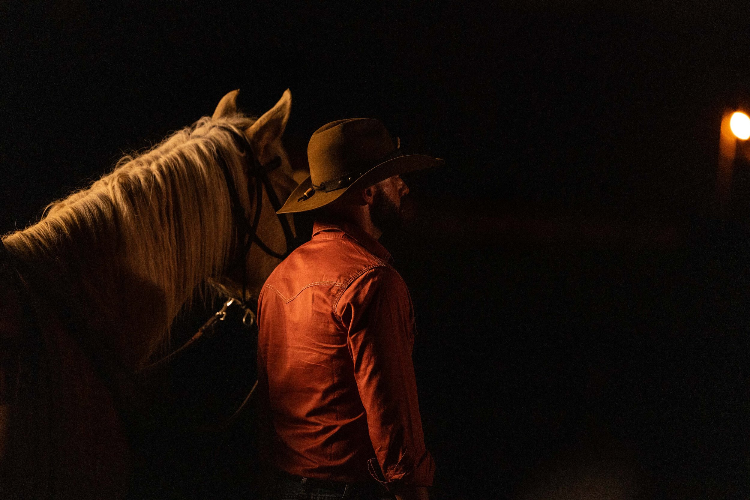 A man with a beard wearing a wide-brimmed hat and a reddish-orange shirt stands next to a horse at night, with a bright light in the distance.