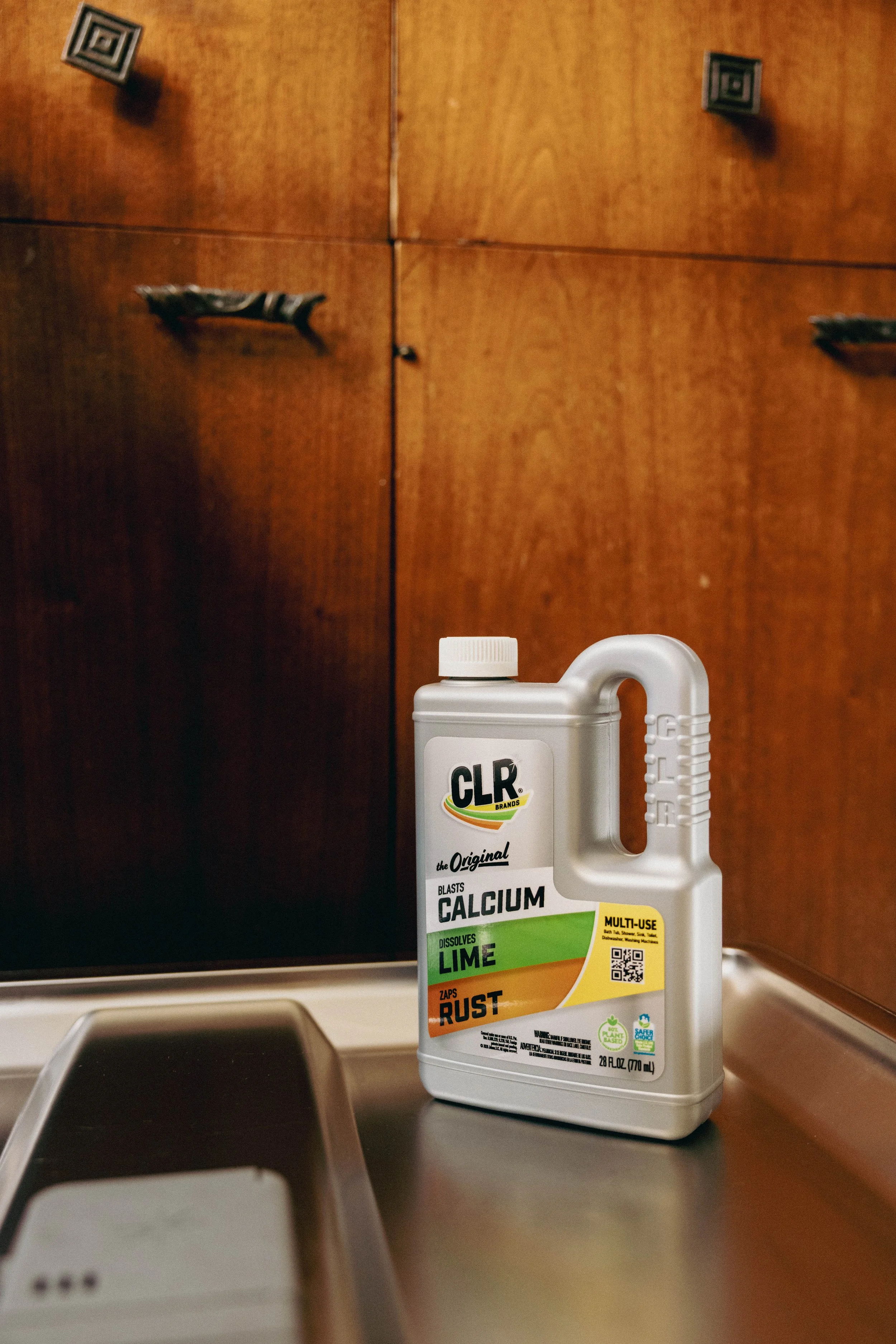 A white bottle of CLR cleaning solution labeled for blasting calcium, dissolving lime, and zapping rust, placed on a kitchen countertop next to a sink with a wooden cabinet in the background.