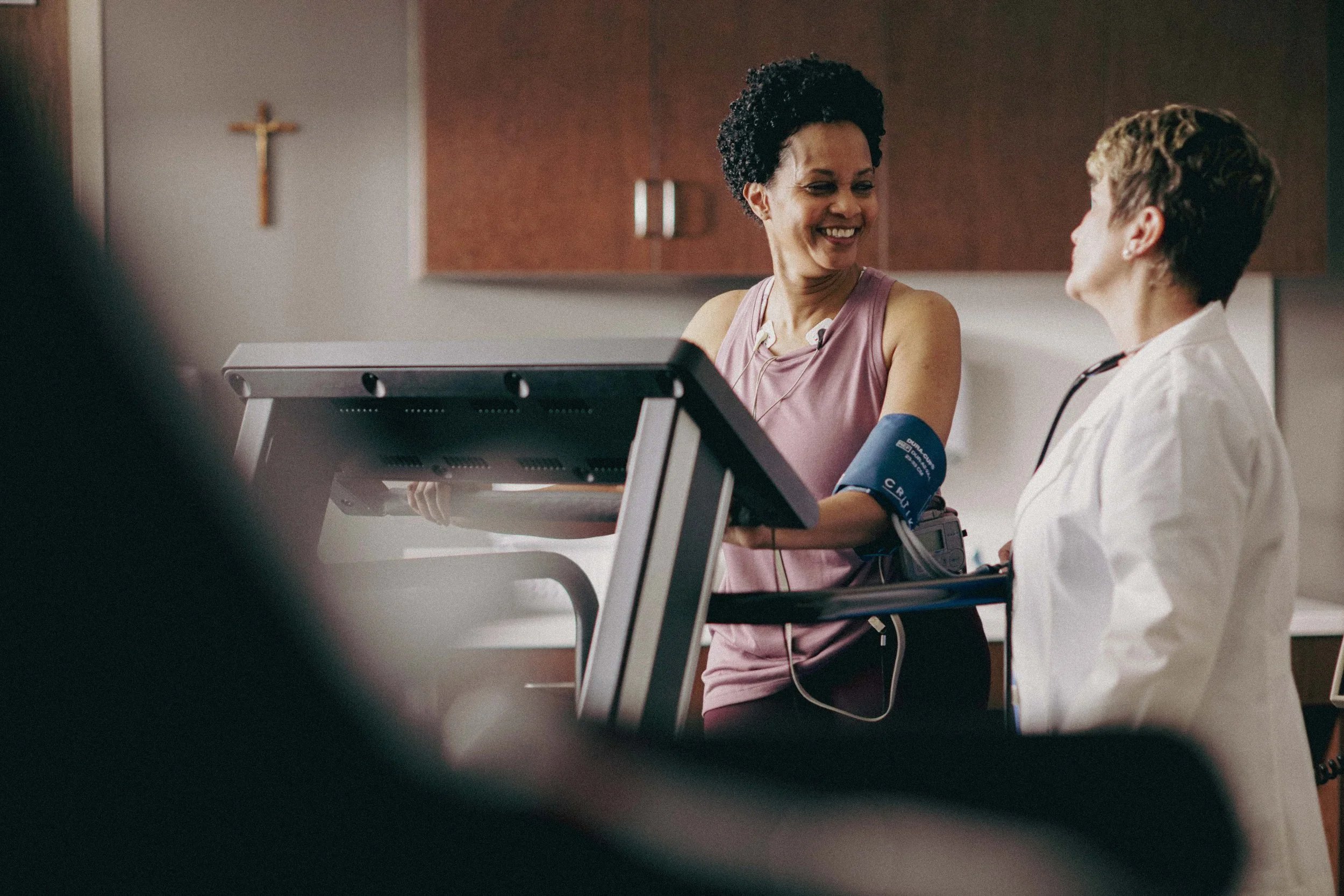 A woman in a pink hospital gown is smiling and talking to a healthcare professional in a white coat, who is listening with a stethoscope. The woman is getting her blood pressure taken in a hospital room with a cross on the wall.