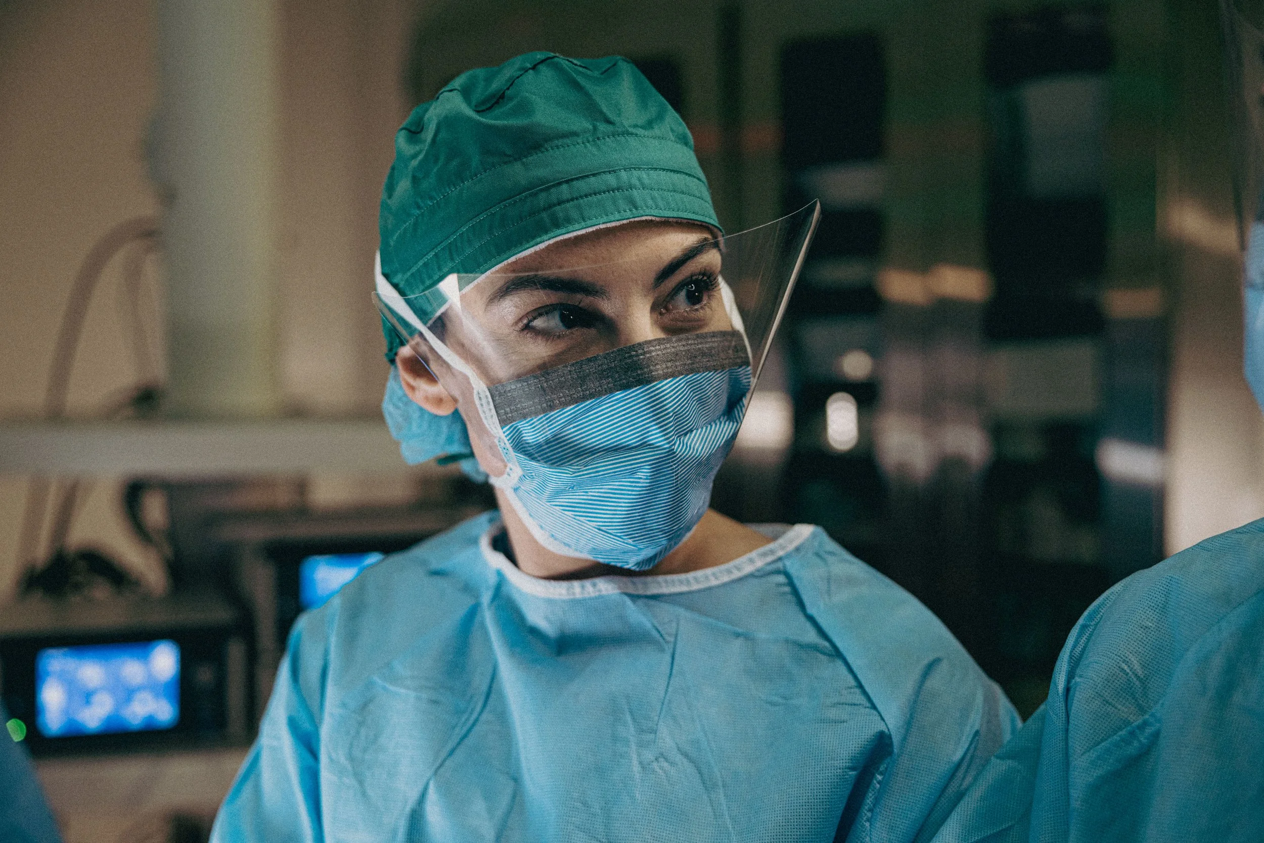A female healthcare worker wearing a green surgical cap, face shield, blue face mask, and scrubs during a medical procedure in an operating room.
