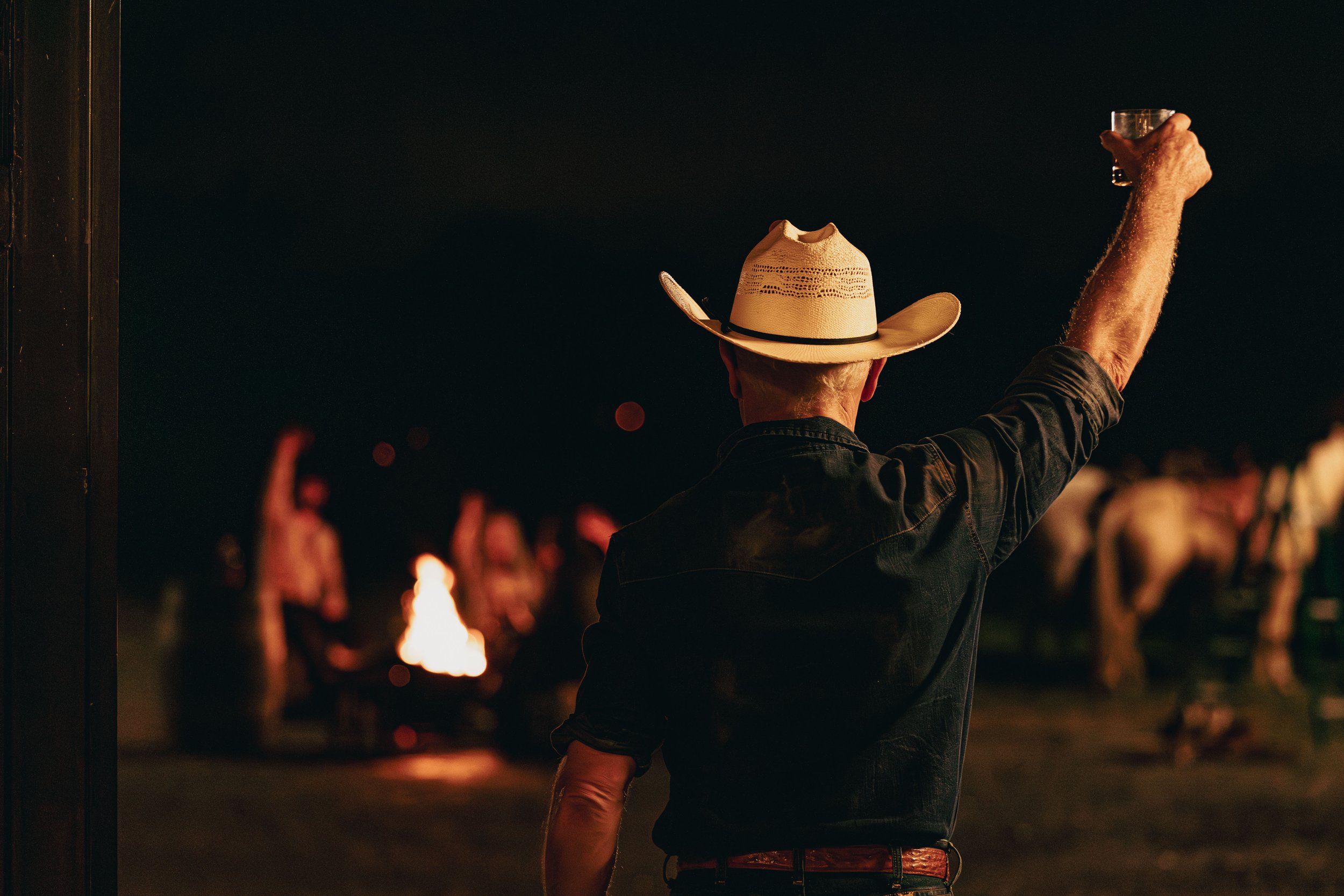 Man wearing a cowboy hat raising a glass at night near a campfire with other people in the background.
