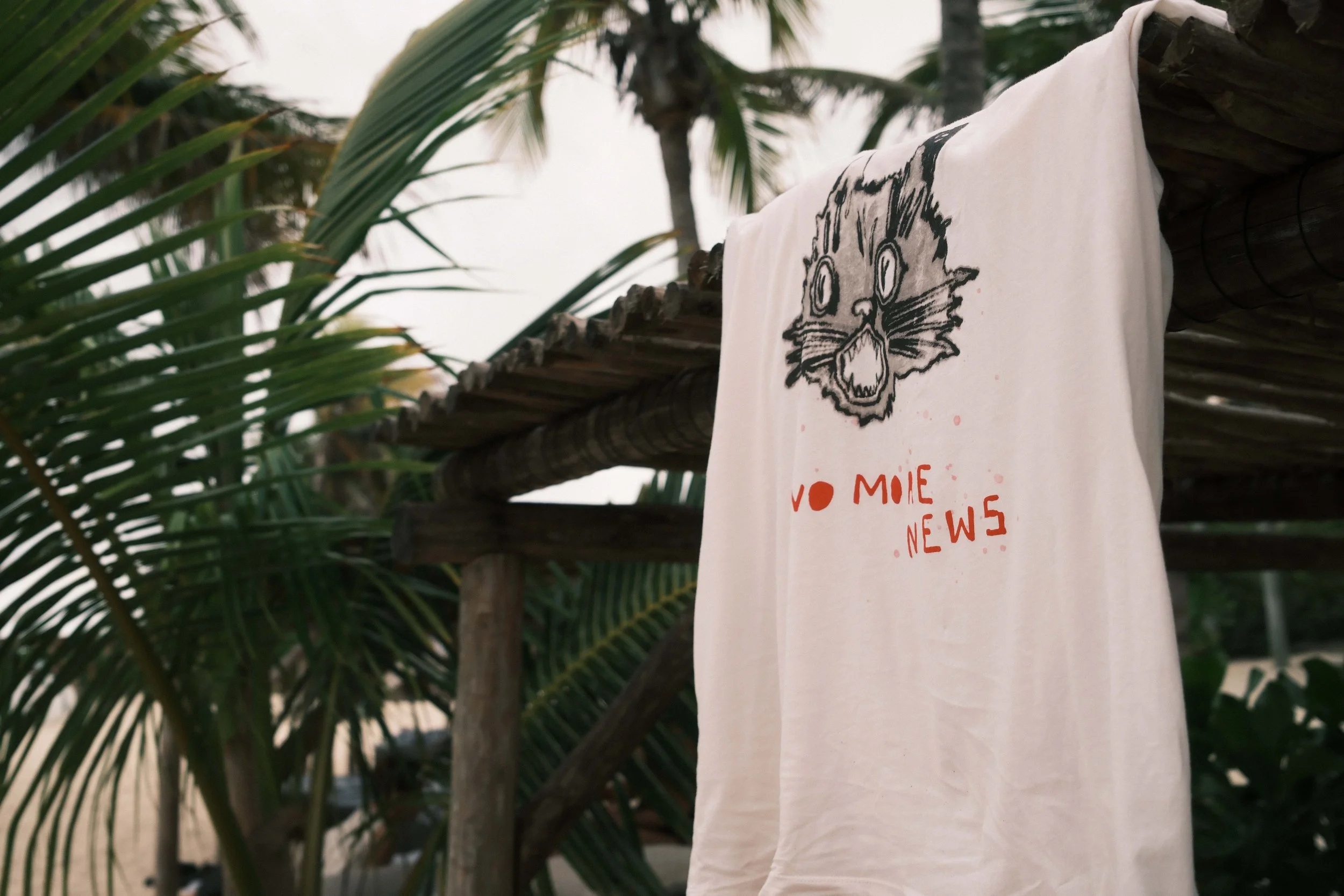 A white T-shirt hanging on a wooden structure outdoors, featuring a black sketch of an animal face and red text that reads 'NO MICE NEWS'. There are green palm leaves surrounding the shirt.