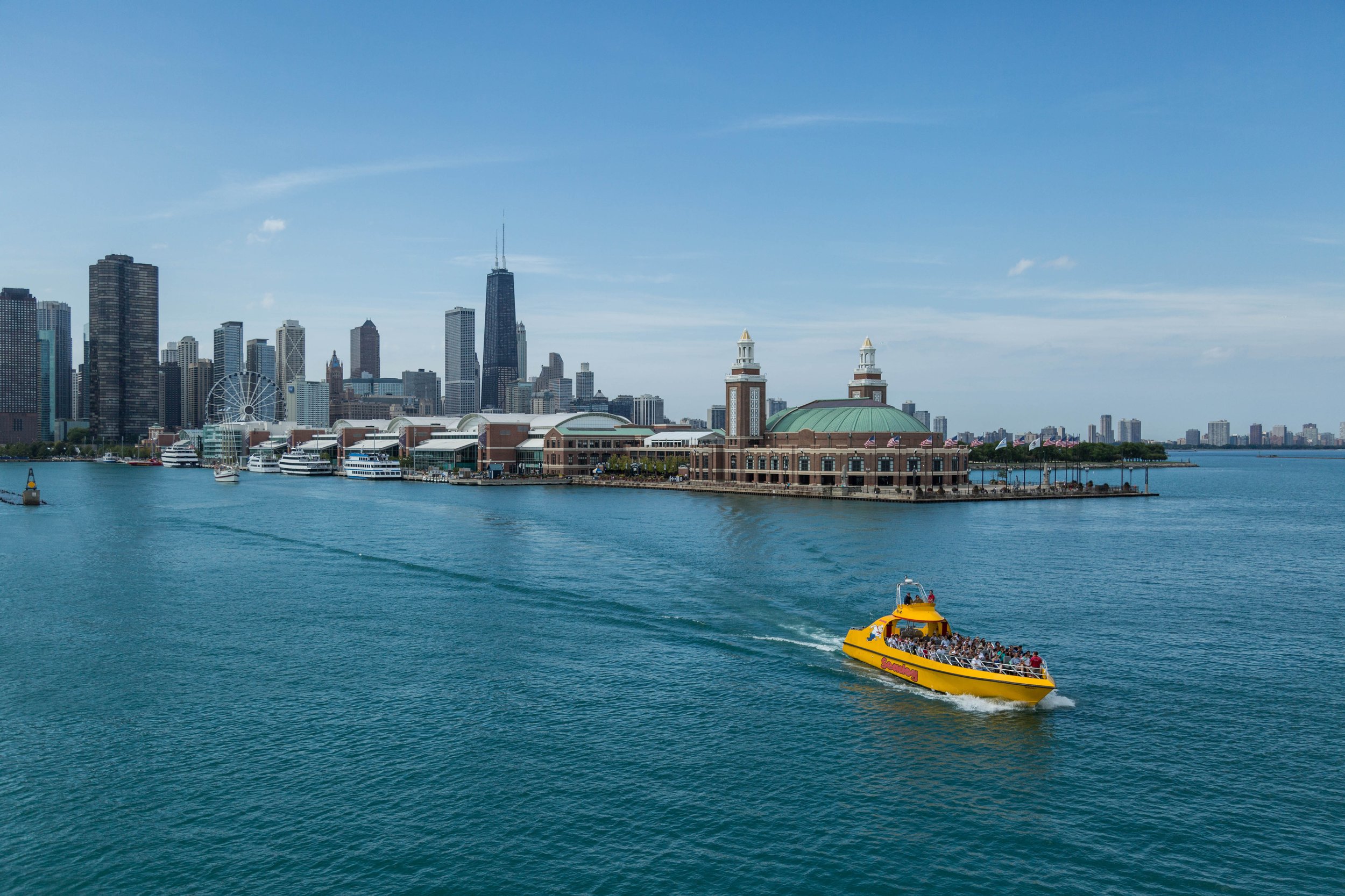 A yellow boat with passengers on Lake Michigan near Navy Pier in Chicago, with the Chicago skyline in the background, including the John Hancock Center and other skyscrapers.
