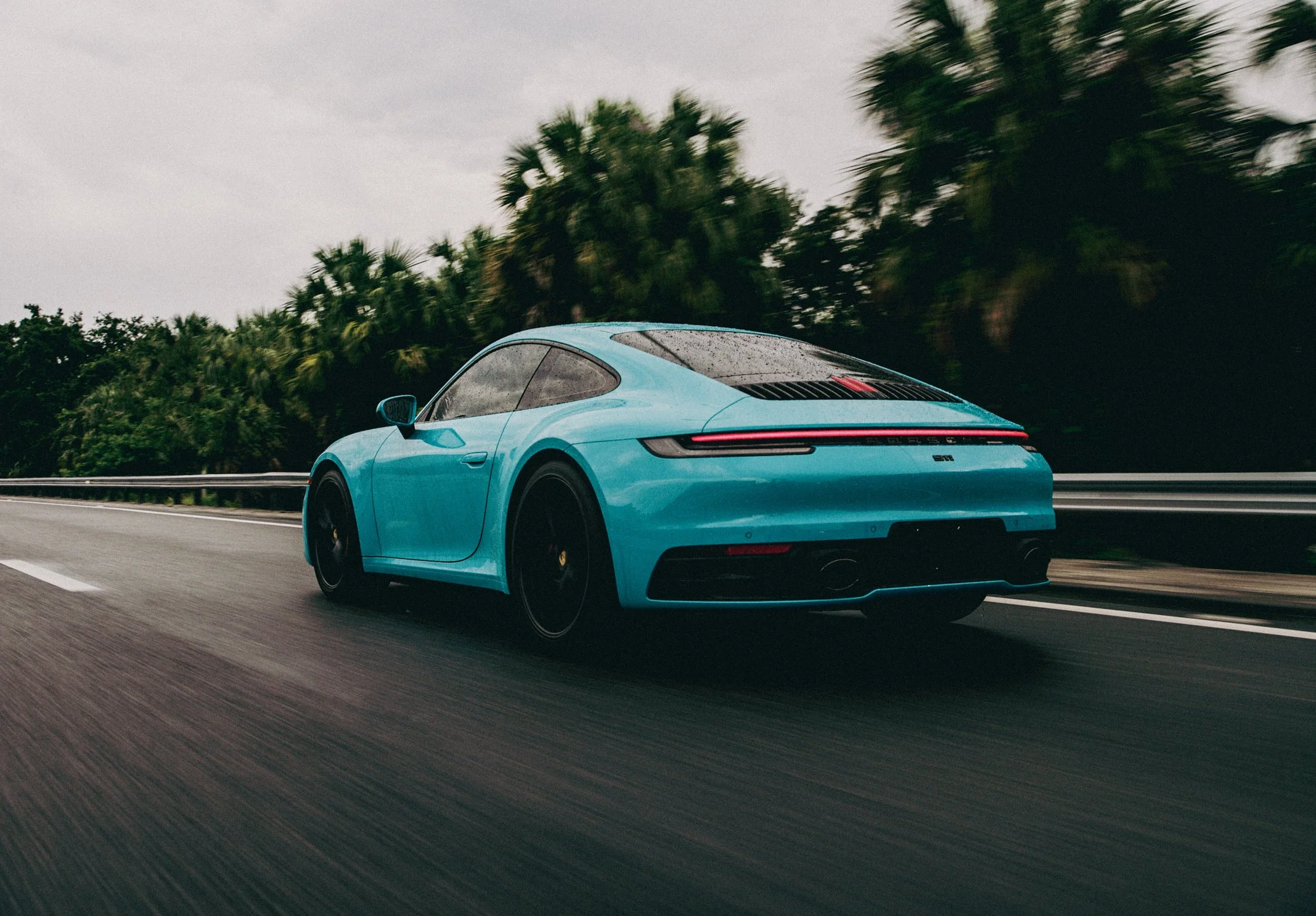 A blue sports car driving on a highway with green trees in the background on a cloudy day.