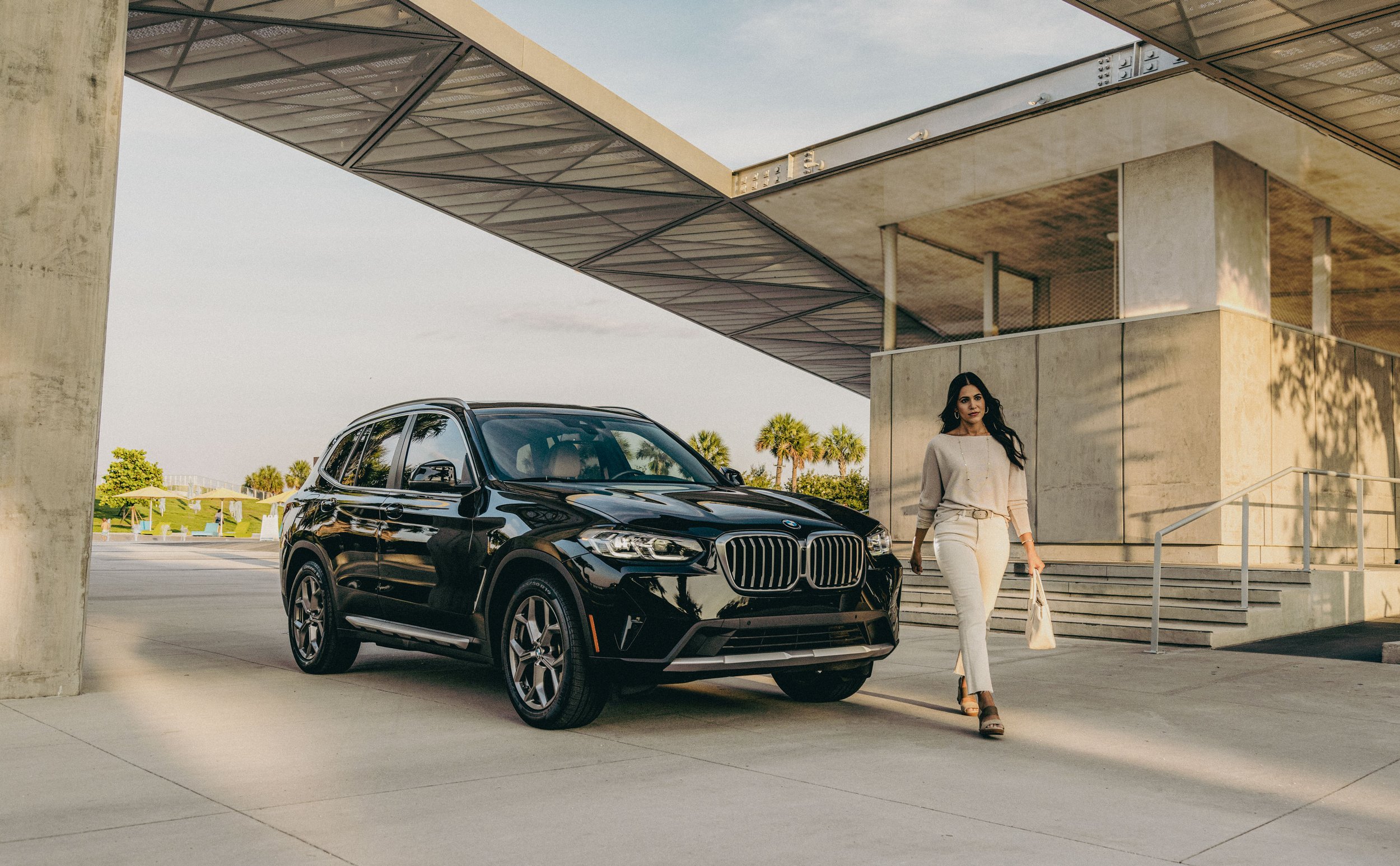 A woman dressed in a beige outfit walking on a sidewalk next to a parked black BMW SUV under a modern building with palm trees in the background.