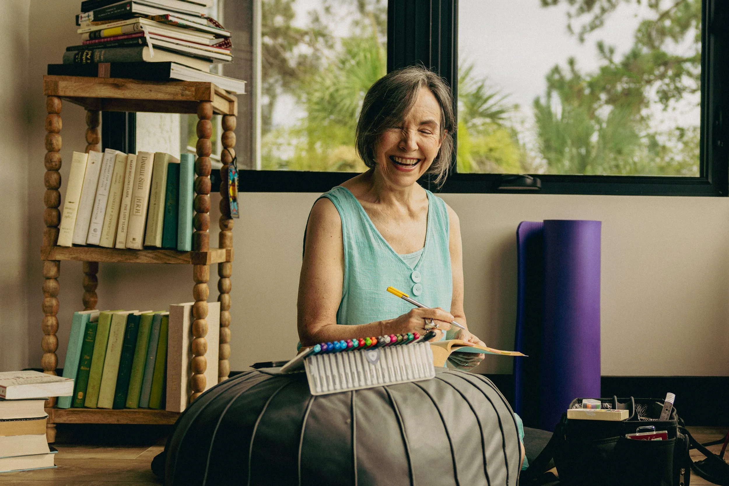 Older woman sitting on a black yoga bolster, smiling, holding a yellow pen and notepad. She has gray hair, wearing a sleeveless turquoise top. There are bookshelves with books behind her, a window with trees outside, and yoga props like a purple foam