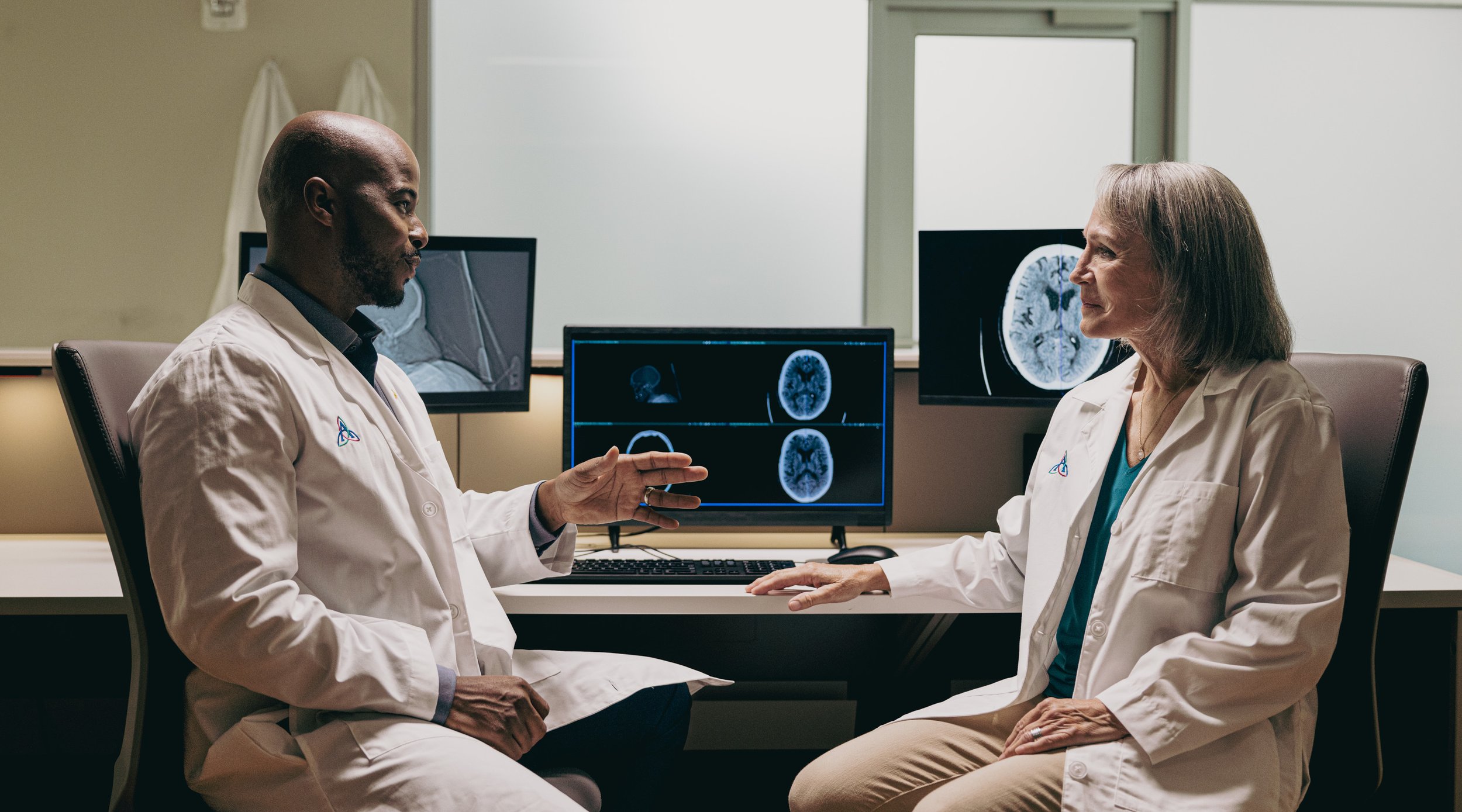 Two doctors, a male and a female, having a discussion in a medical office with brain scan images on the computer monitors behind them.
