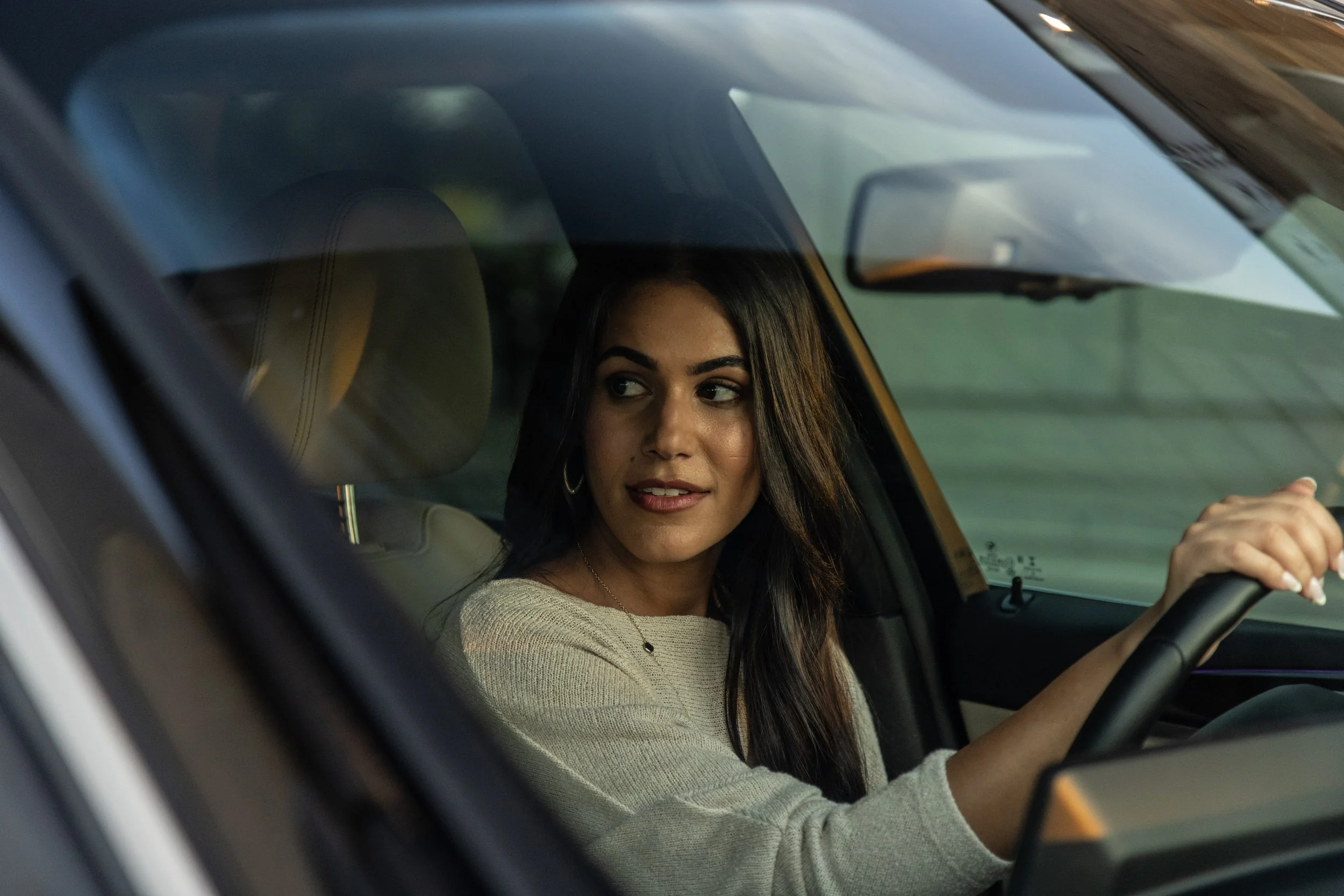 Woman with dark hair driving a car, looking to the side, wearing a light-colored sweater.