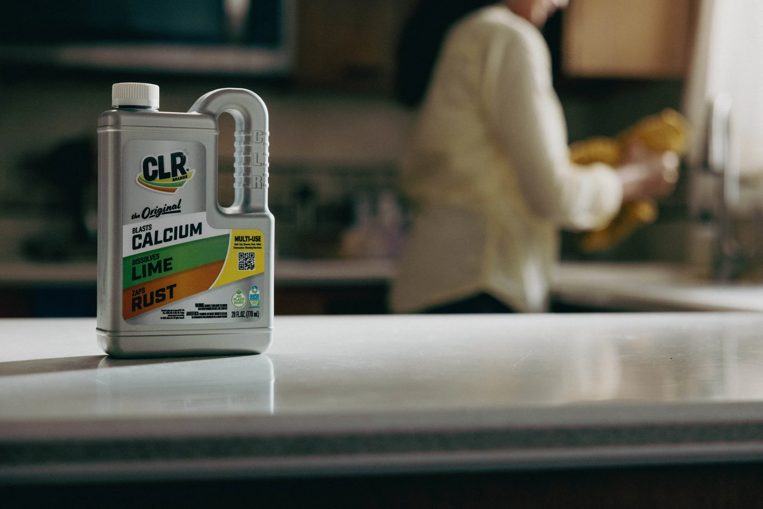 A bottle of CLR cleaner on a kitchen counter with a person in the background washing dishes.