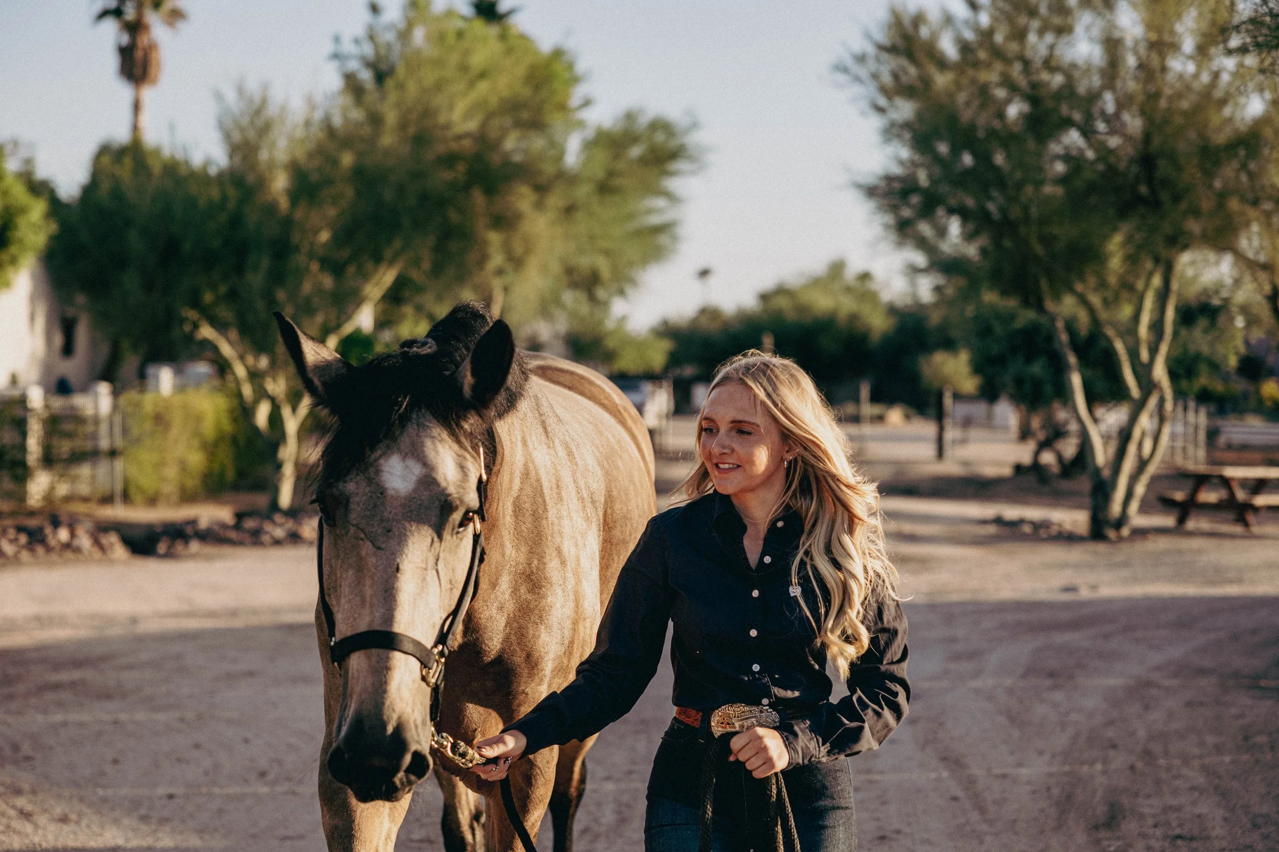 A woman walking a light brown and black horse on a path in a park during the daytime with trees and clear sky in the background.