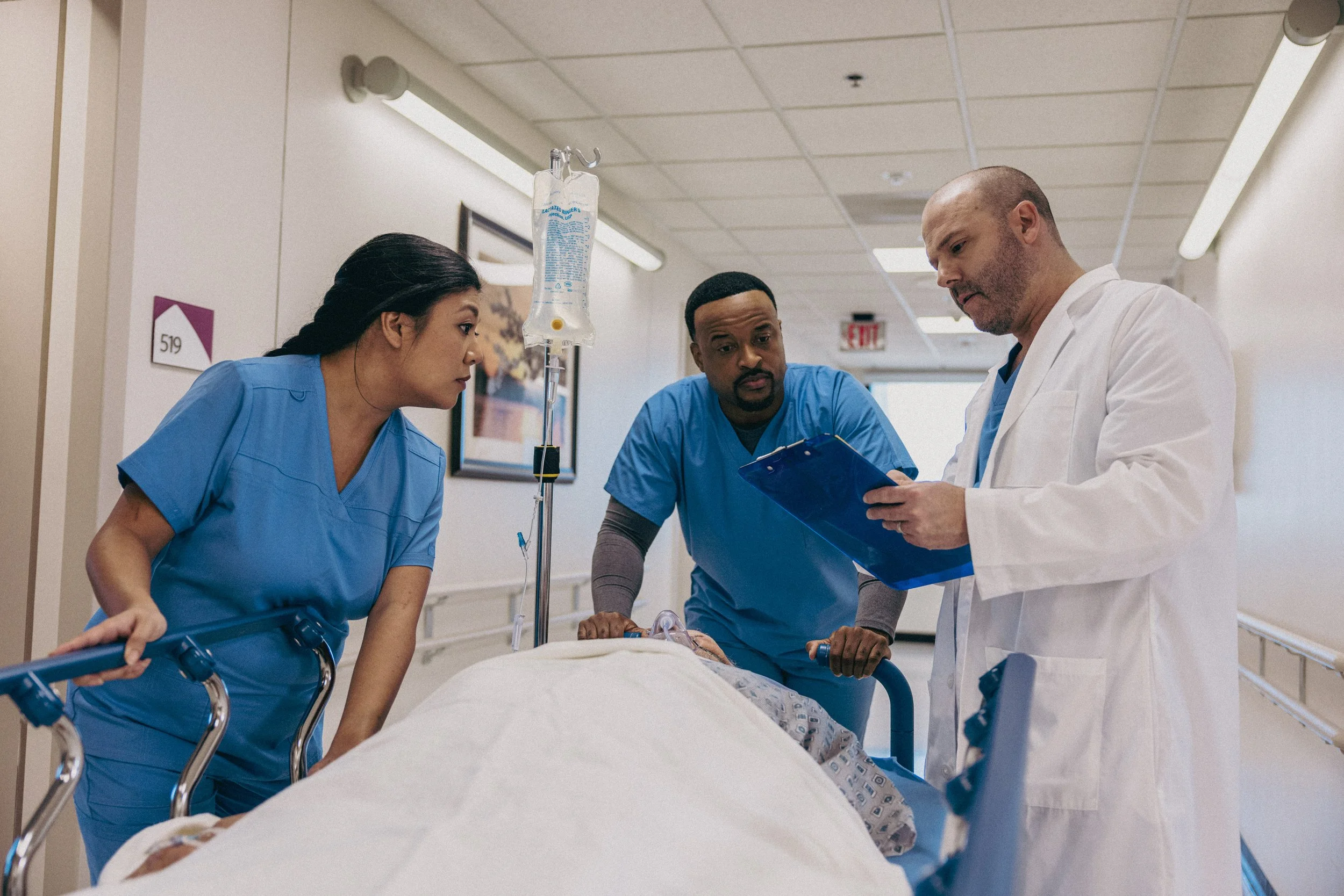 Three medical professionals, two women in blue scrubs and one man in a white coat, are gathered around a hospital bed with a patient, reviewing medical charts and discussing treatment.