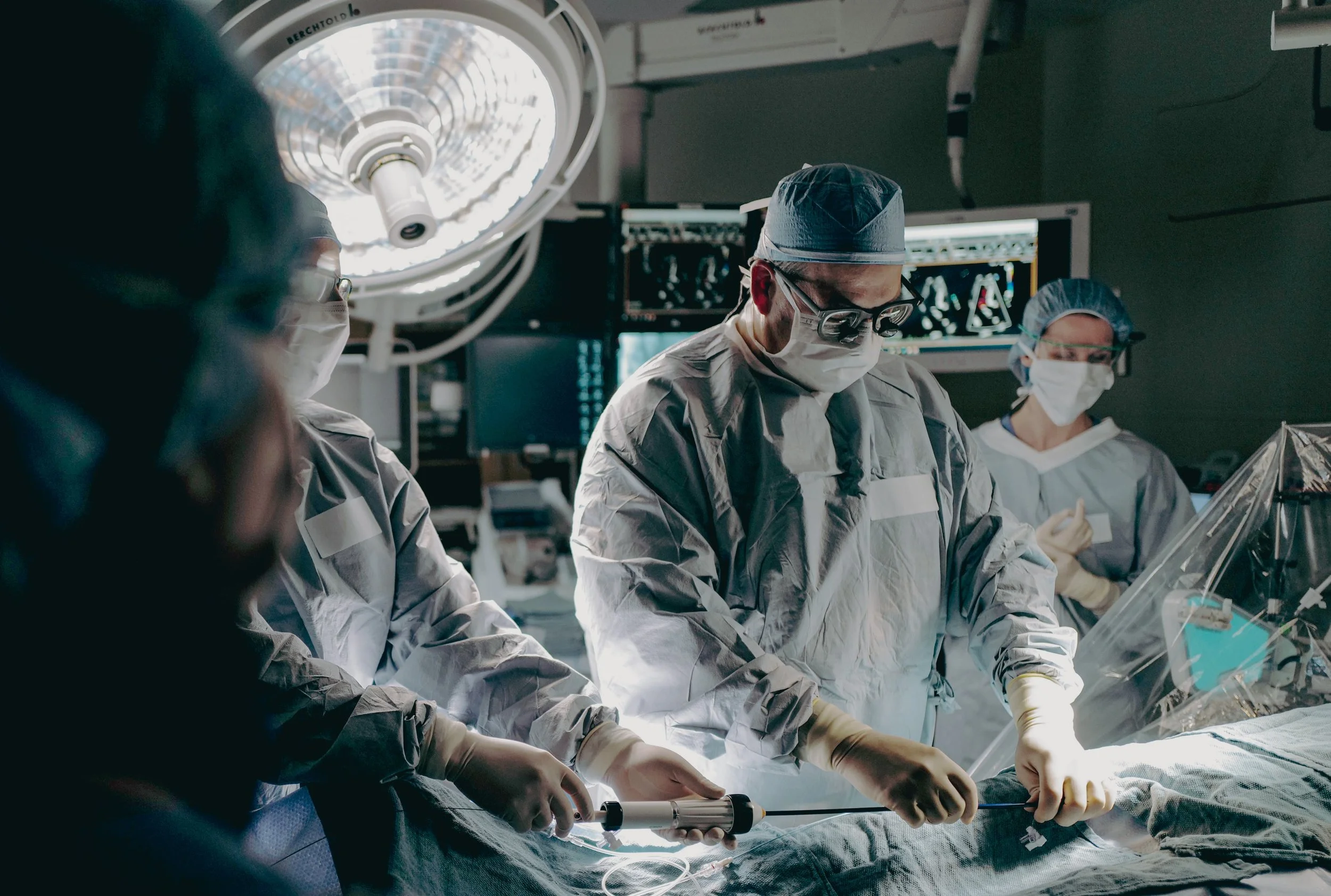 Medical team performing surgery in an operating room, wearing surgical masks, gowns, and gloves, with monitors and surgical lights overhead.