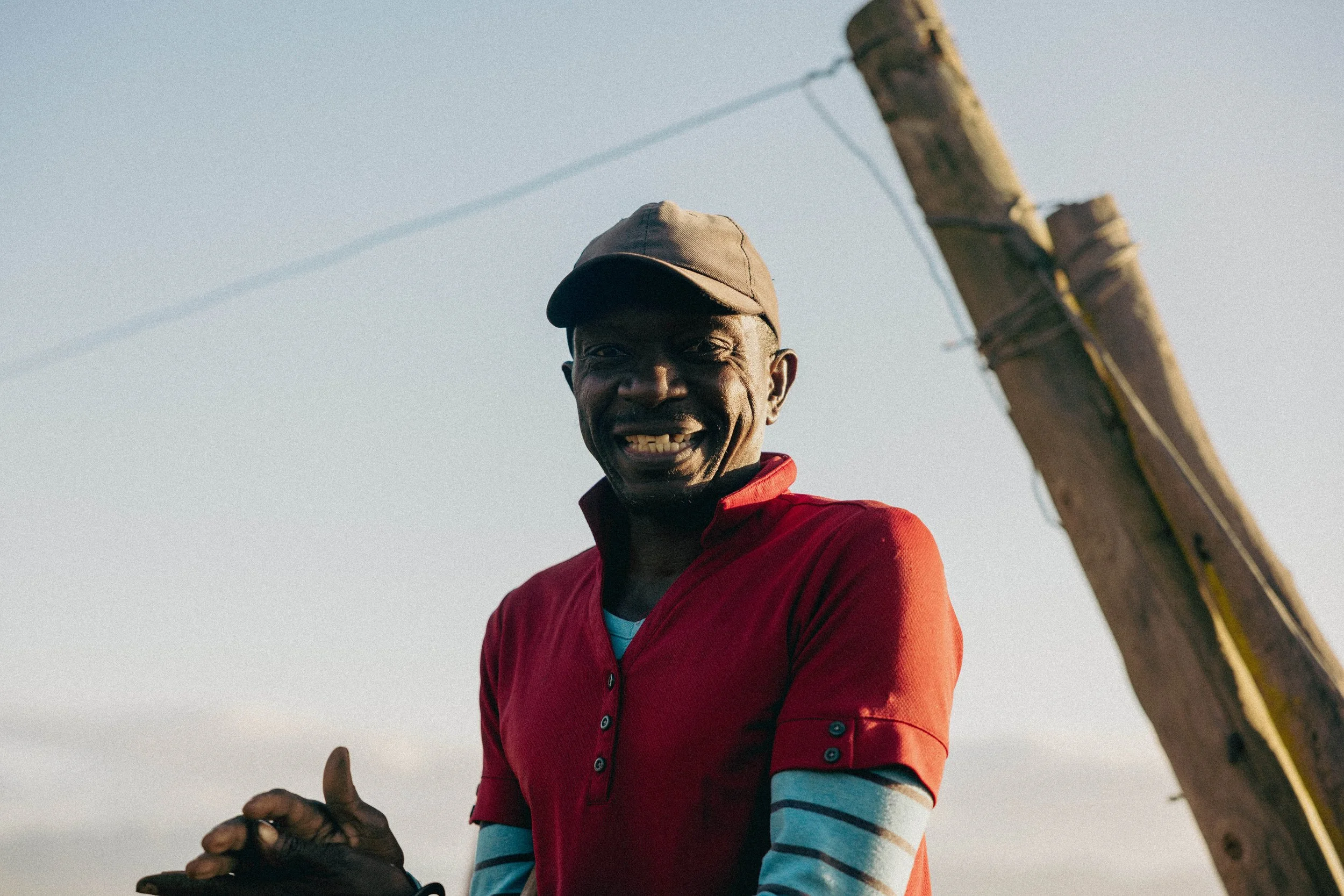 A smiling man wearing a red shirt with a striped undershirt, a brown cap, standing outdoors near a wooden utility pole against a clear sky.