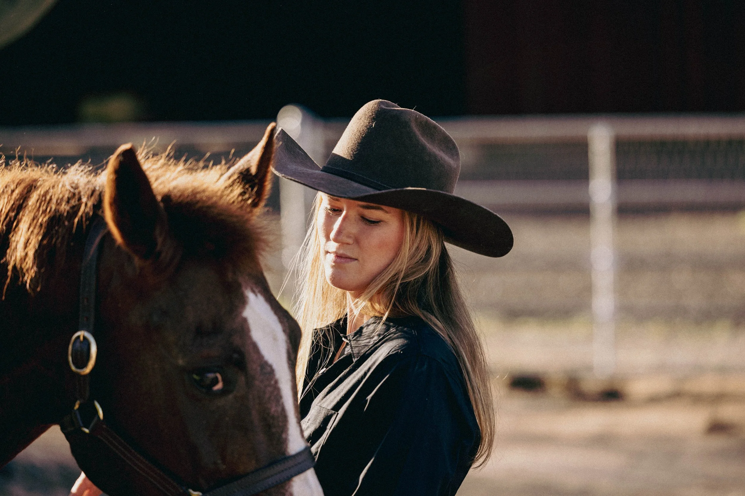 A woman wearing a wide-brimmed hat and black shirt standing next to a horse on a farm or ranch.
