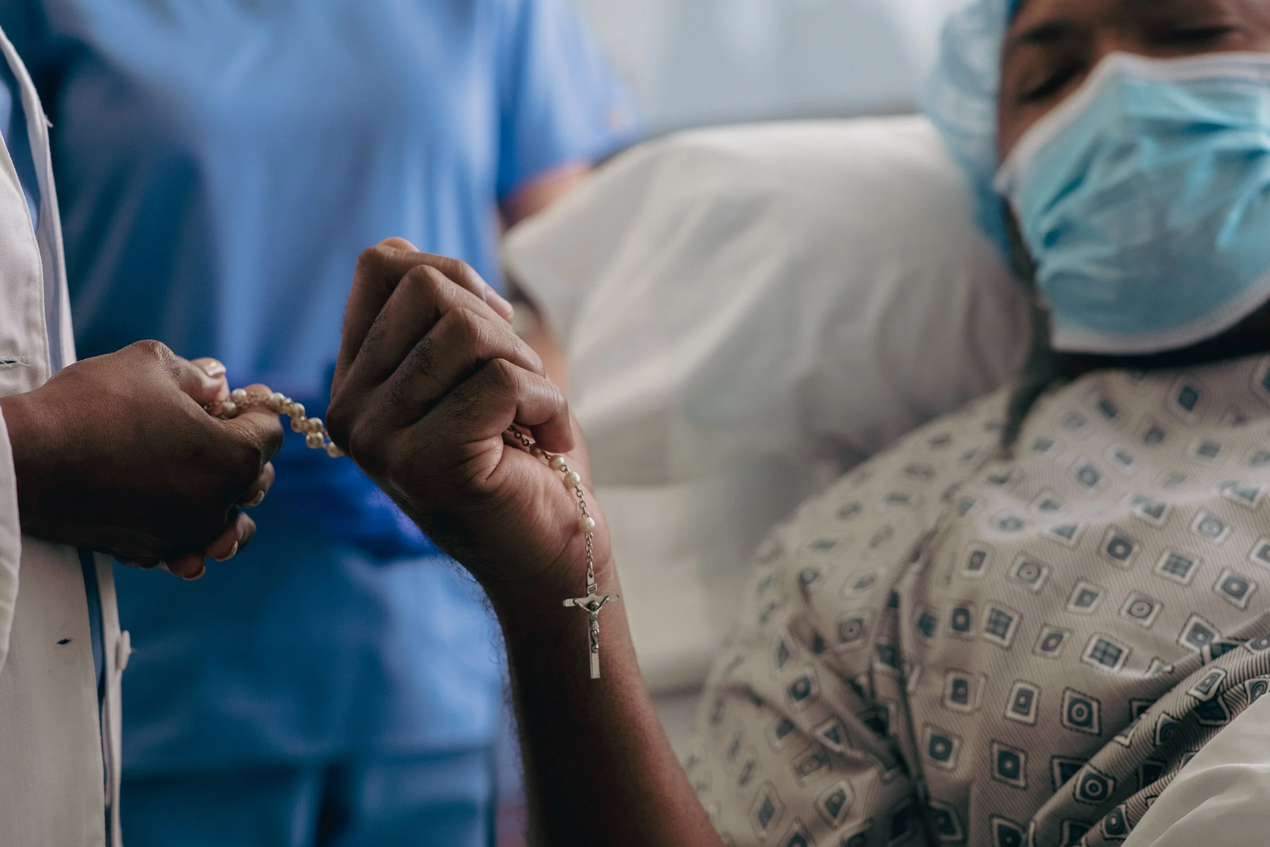 A person in a hospital bed holding a rosary with a crucifix, wearing a hospital gown and a face mask, with another person in medical scrubs holding their hand.