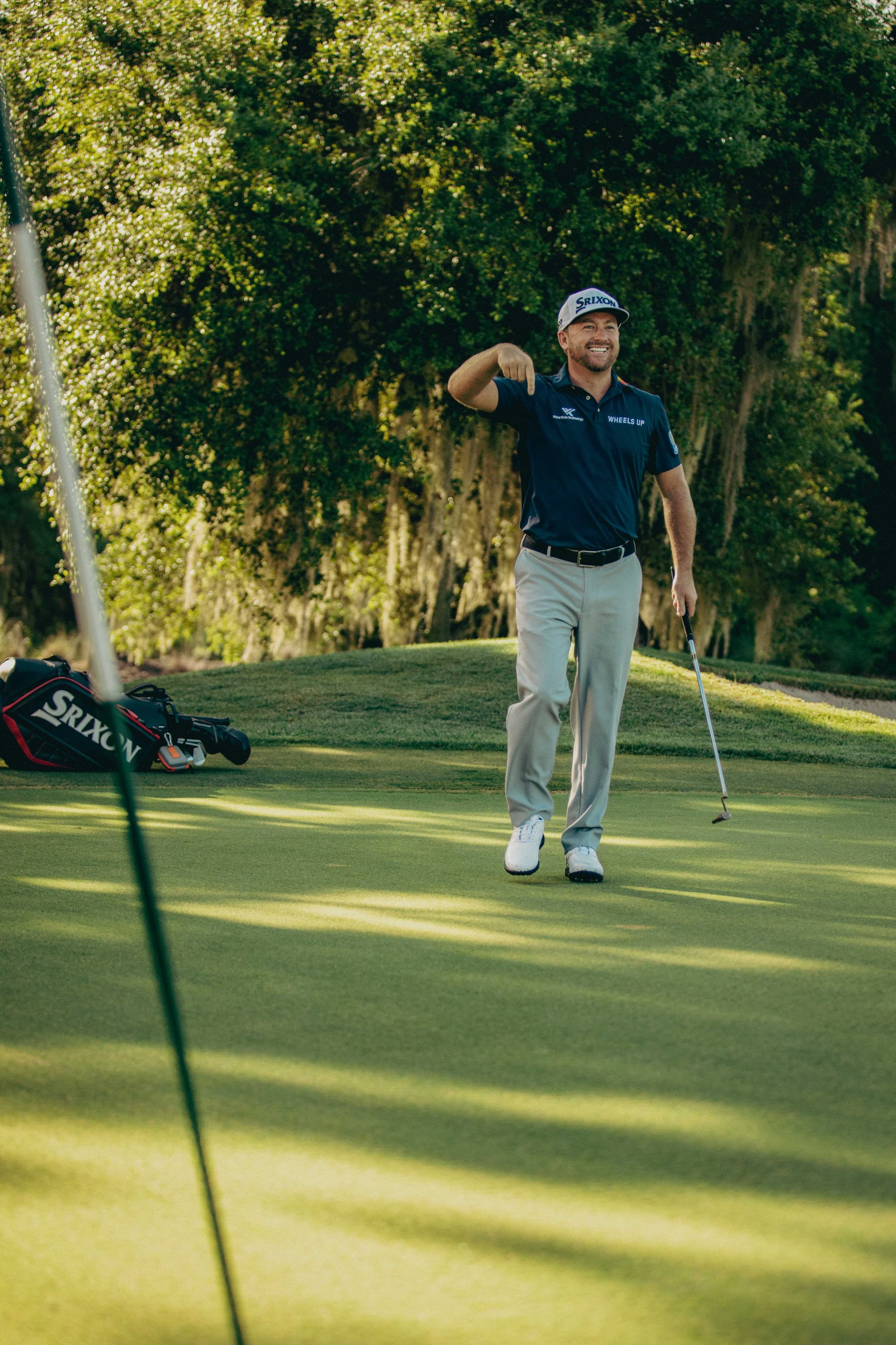 A man smiling and waving on a golf course, holding a golf club, with a golf bag nearby, surrounded by trees.