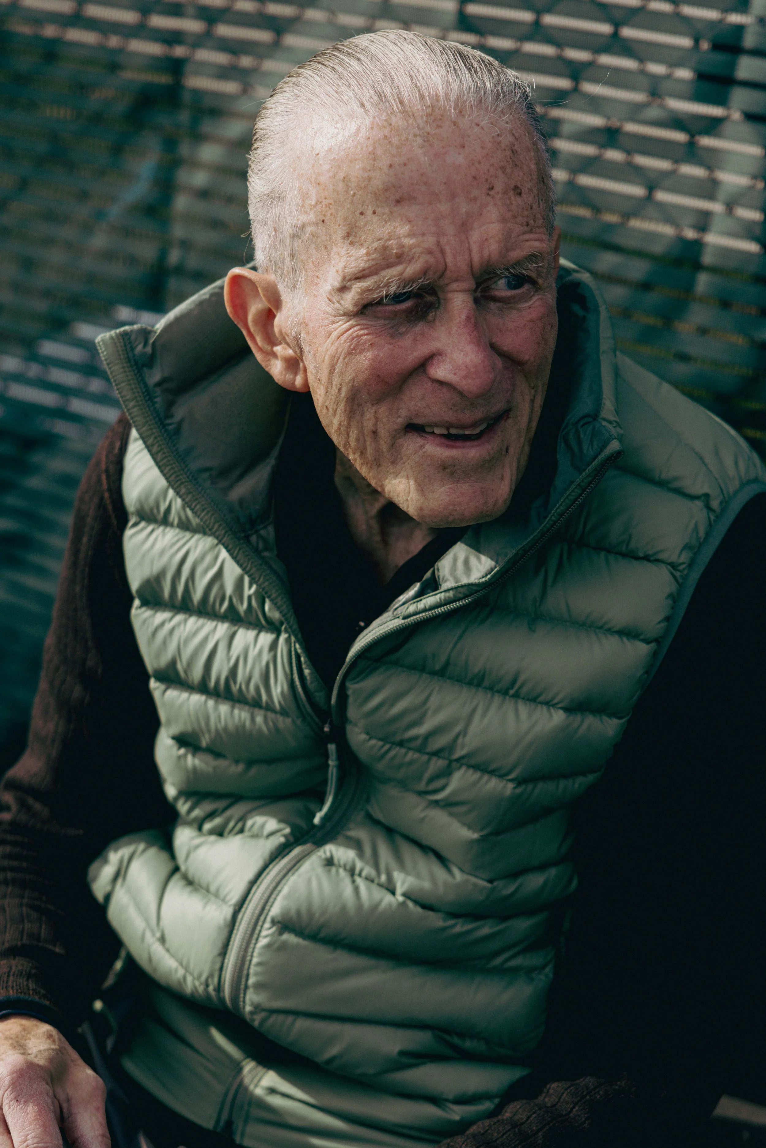 An elderly man with a distressed or intense facial expression, wearing a light-colored puffer vest and a dark long-sleeve shirt, in front of a metal fence.