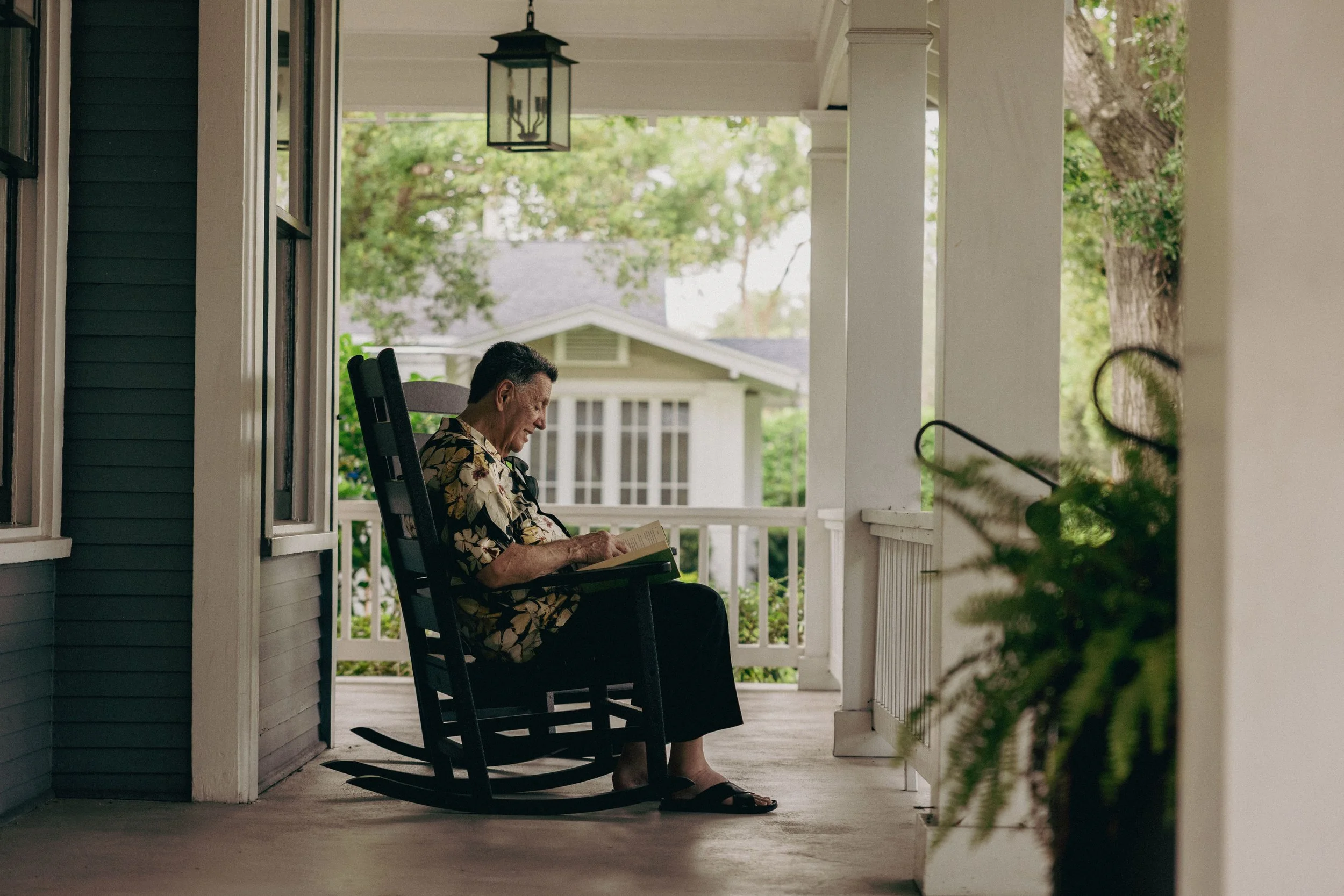 An elderly man sits on a porch in a rocking chair, reading a book and smiling, surrounded by greenery and a white railing.