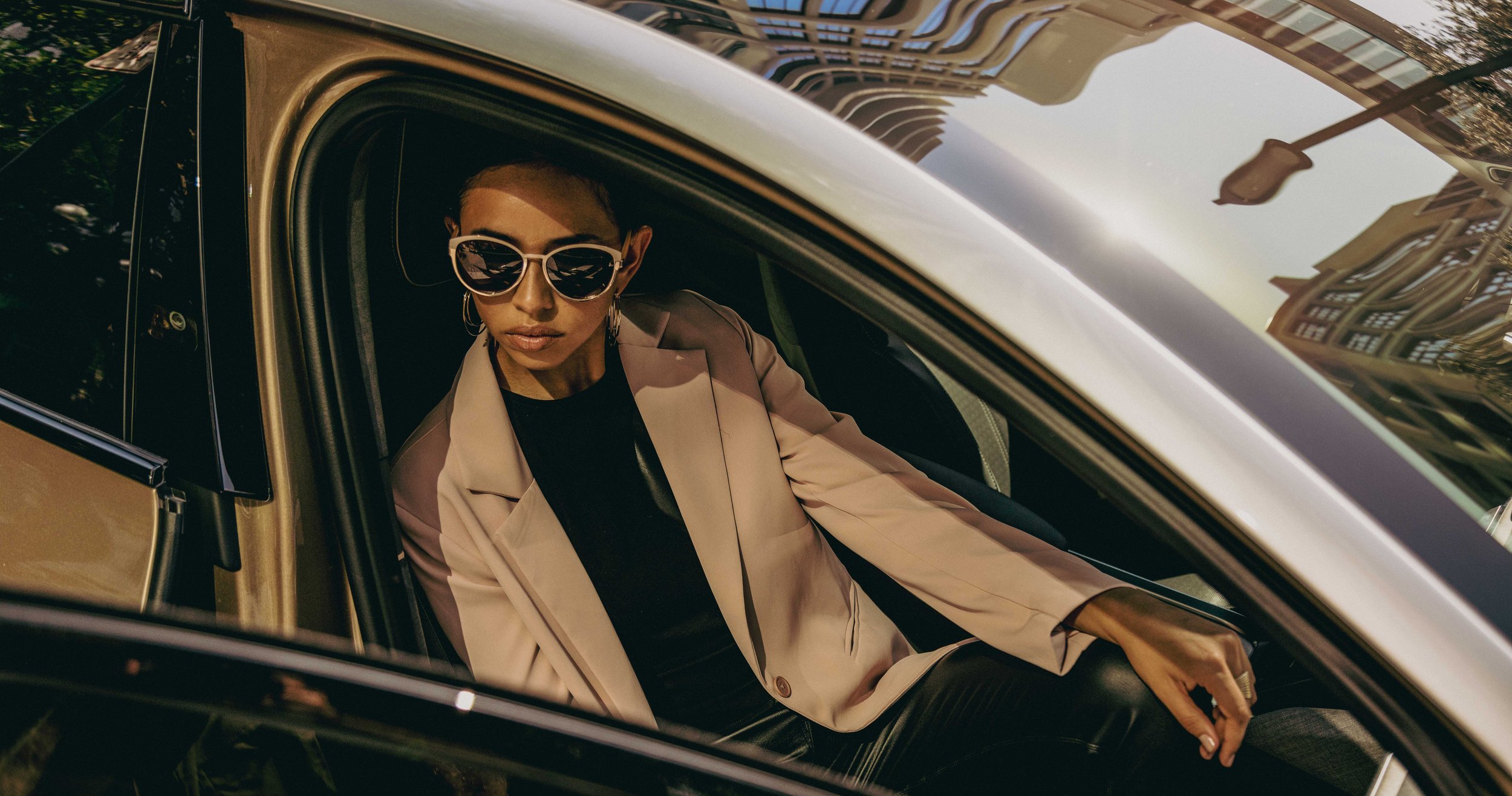 A woman sitting in the driver's seat of a black car, wearing sunglasses, a beige blazer, black top, and hoop earrings, with a city building reflected on the car window.
