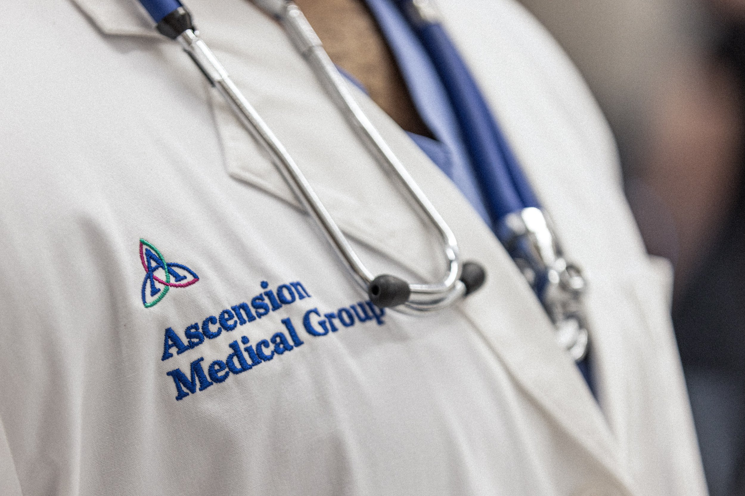 Close-up of a person wearing a white medical coat with an Ascension Medical Group logo and a stethoscope hanging around their neck.