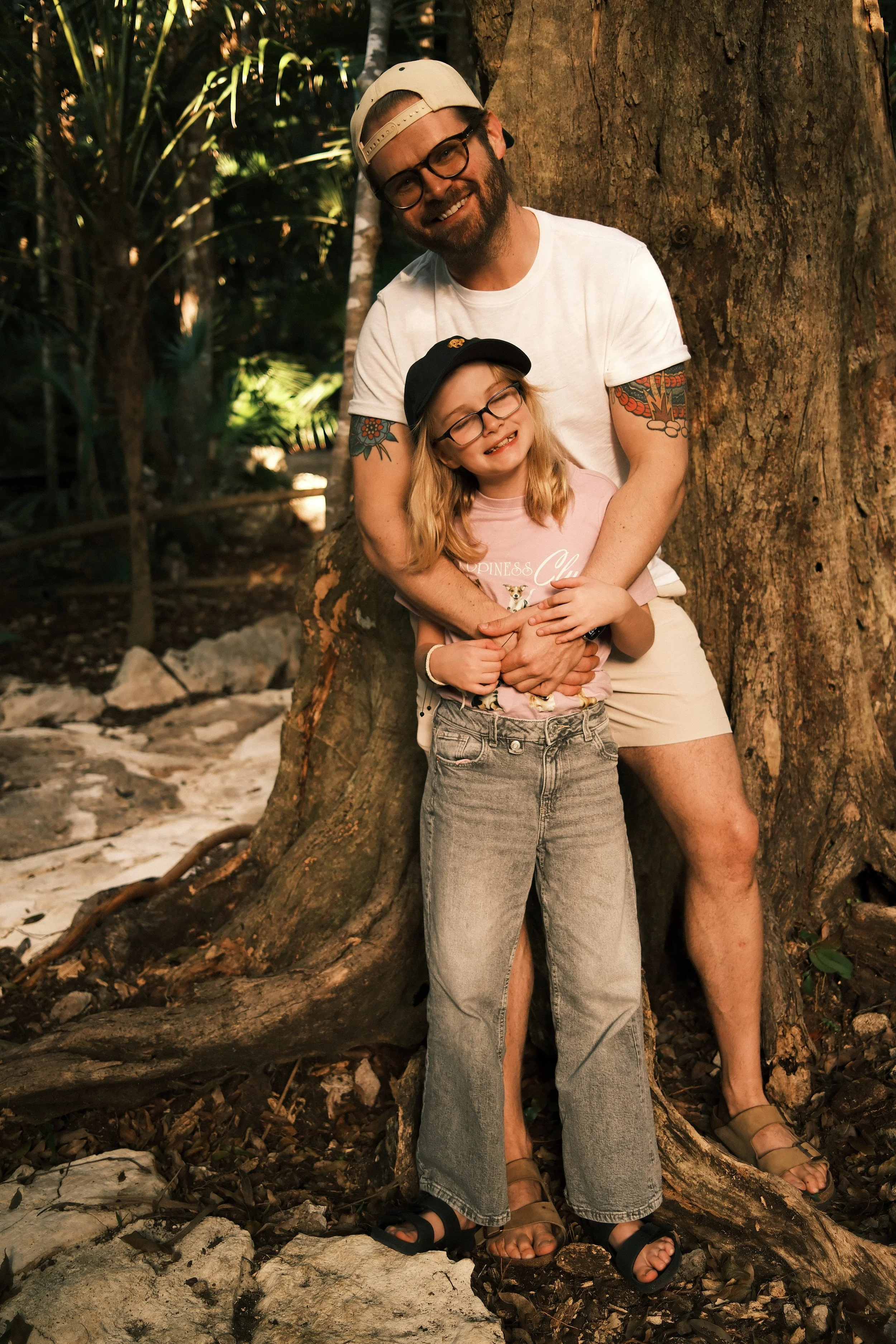 A man with a beard, glasses, tattoos, wearing a cap backward, white t-shirt, khaki shorts, and sandals, hugging a young girl with glasses, long blonde hair, wearing a black cap, pink t-shirt, jeans, and sandals, standing in front of a large tree in a jungle setting.