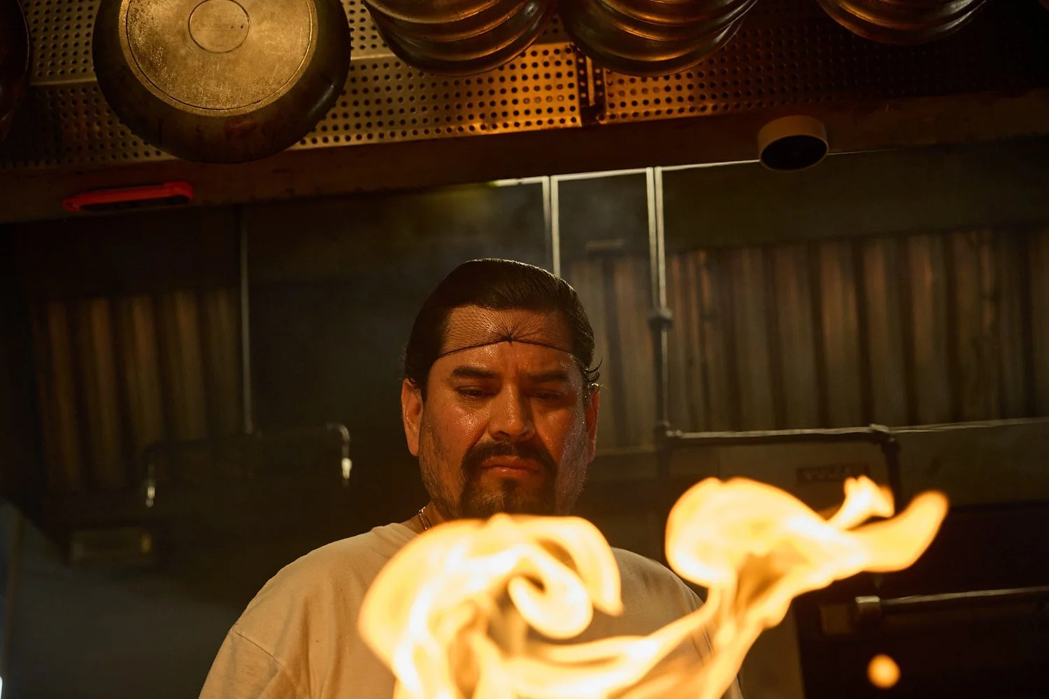A man with dark slicked-back hair and a beard, looking down with a serious expression, is standing in front of a fire, in what appears to be a restaurant or kitchen setting.