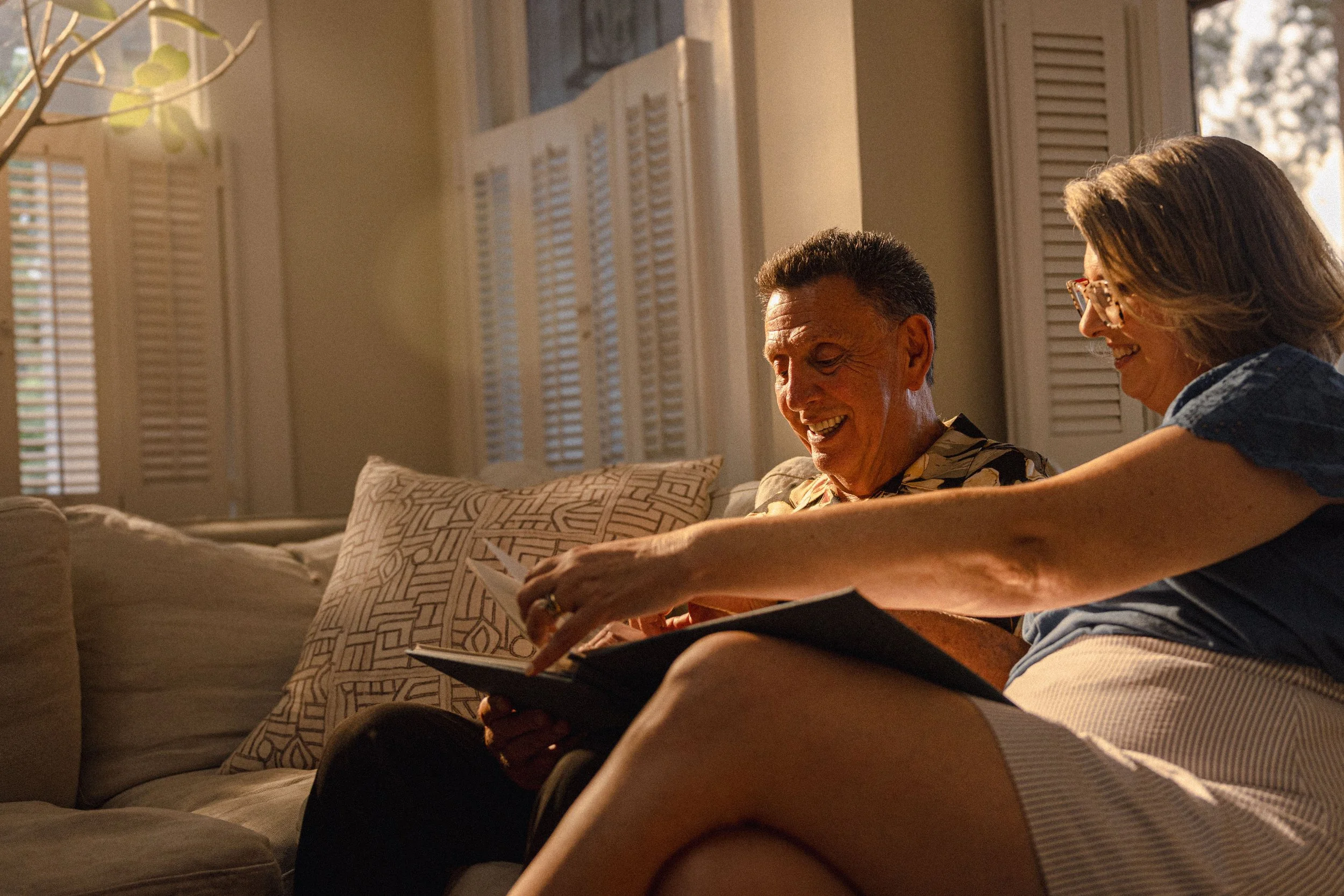 A middle-aged man and woman sitting on a sofa, looking at a photo album together and smiling in a cozy, well-lit living room.