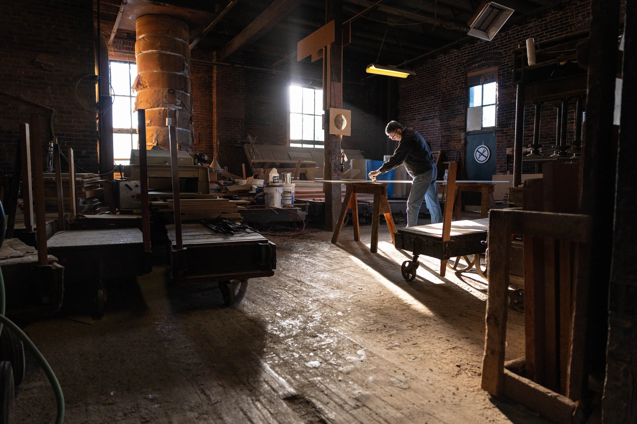 A person working in a woodworking workshop filled with tools, wood, and machinery, illuminated by natural sunlight from large windows.