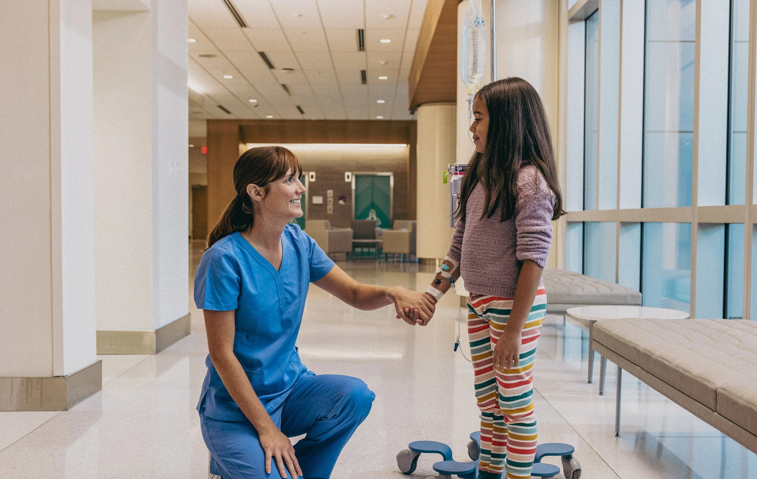 A nurse kneeling and shaking hands with a young girl in a hospital lobby. The girl is standing on a rolling scale, wearing striped leggings and a pink sweater. The nurse is in blue scrubs and the background shows a waiting area with chairs and large 