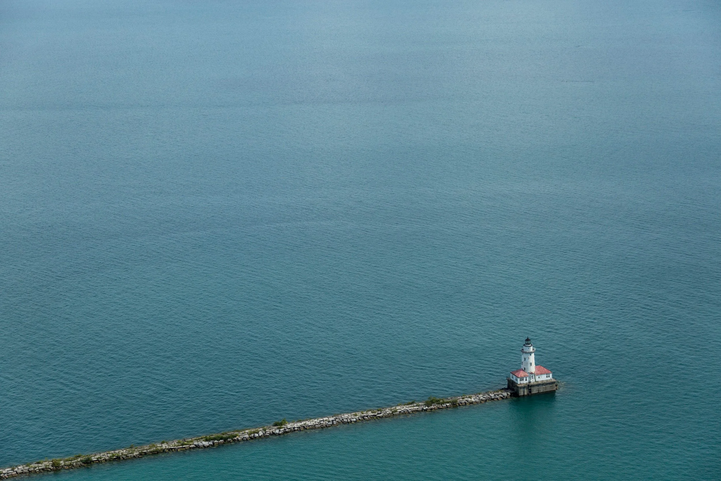 A lighthouse situated at the end of a breakwater extending into a large body of water, with calm blue-green water surrounding it.