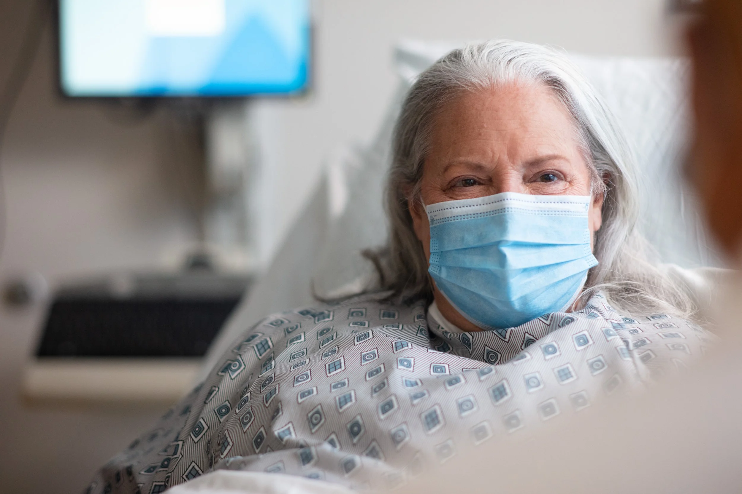 An elderly woman with gray hair wearing a blue face mask and hospital gown, lying in a hospital bed.