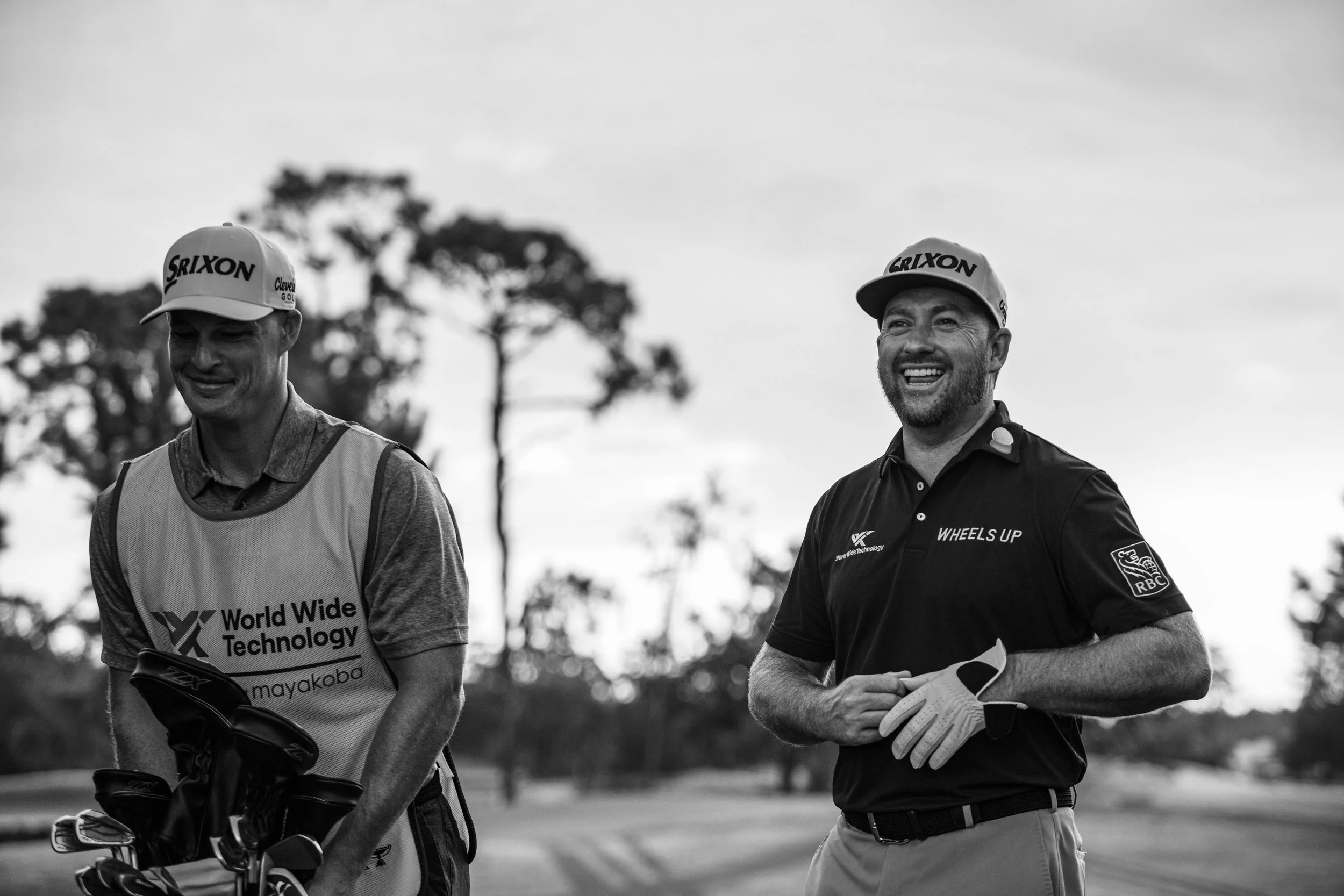 Two smiling men in golf attire, one holding golf clubs, standing outdoors on a golf course with trees in the background.