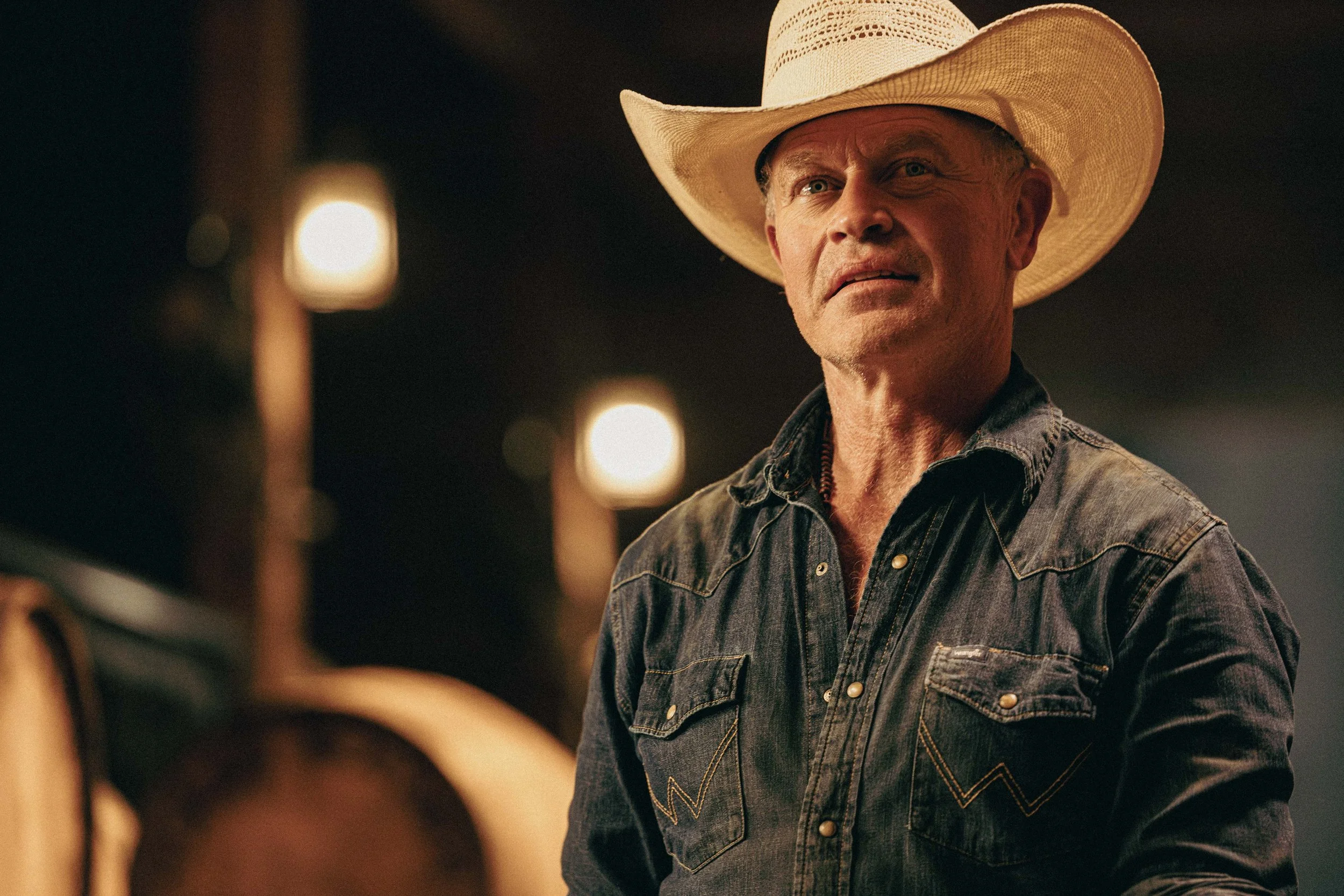 A man wearing a cowboy hat and denim shirt looks to the side against a dark background with warm lighting.