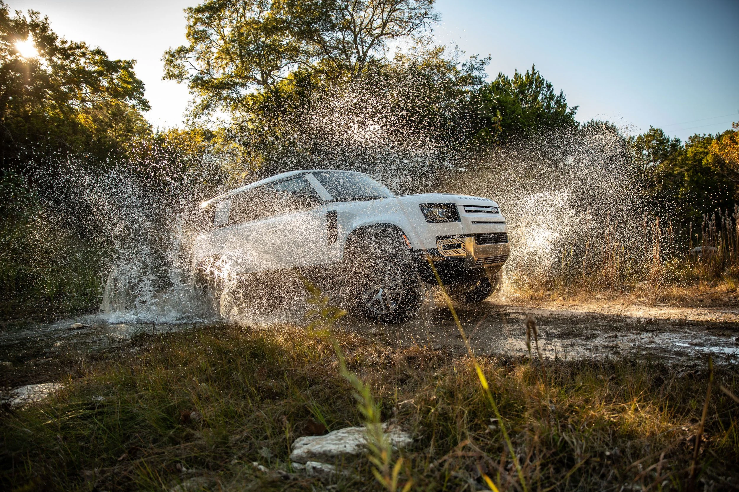 A white off-road vehicle splashing through a muddy puddle on a dirt trail, surrounded by trees and grass with sunlight filtering through the foliage.