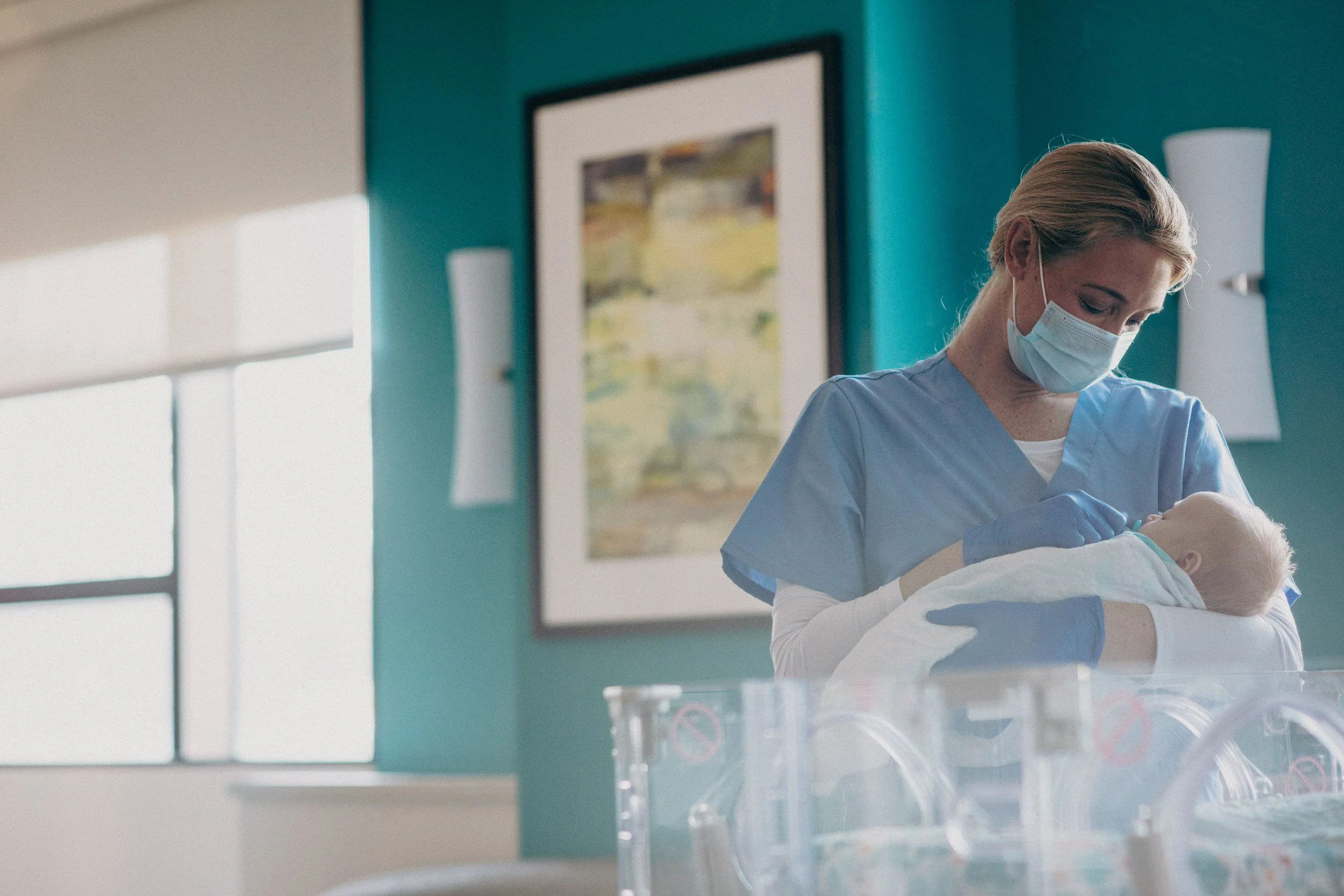 A healthcare worker in blue scrubs, wearing a face mask and gloves, caring for a newborn baby in an incubator in a hospital nursery.