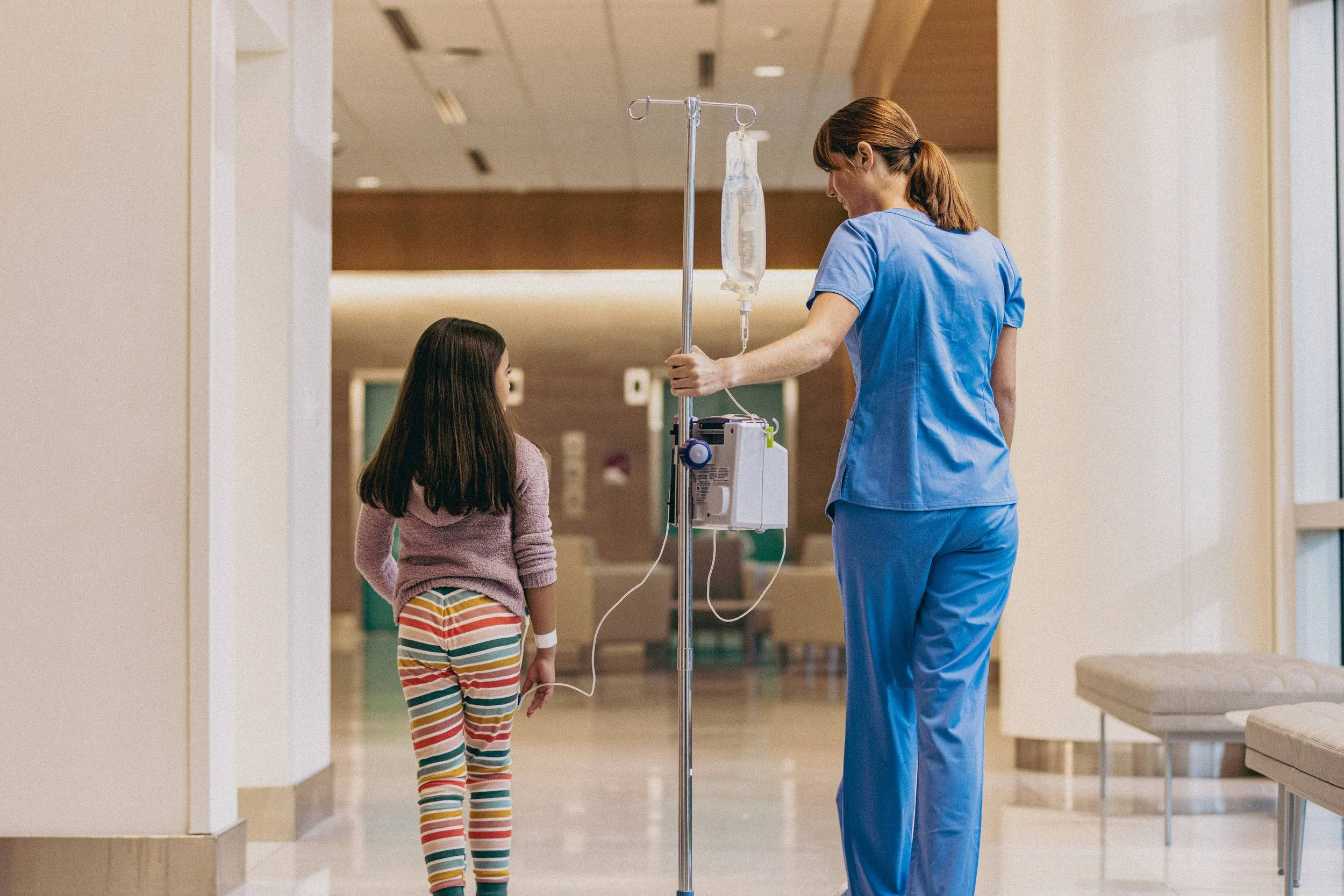 A young girl standing in a hospital corridor, talking to a nurse who is holding an IV stand with medical supplies.